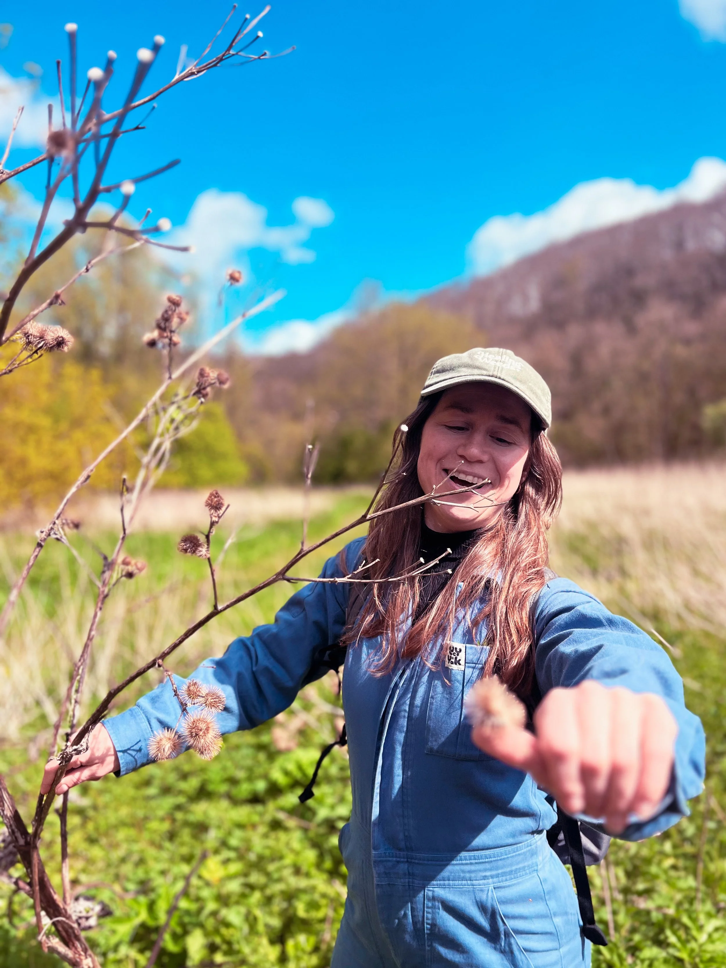 A woman outdoors in a natural setting with mountains and blue sky in the background. She has long brown hair, is wearing a beige cap, a blue jacket, and is smiling while reaching out towards the camera.