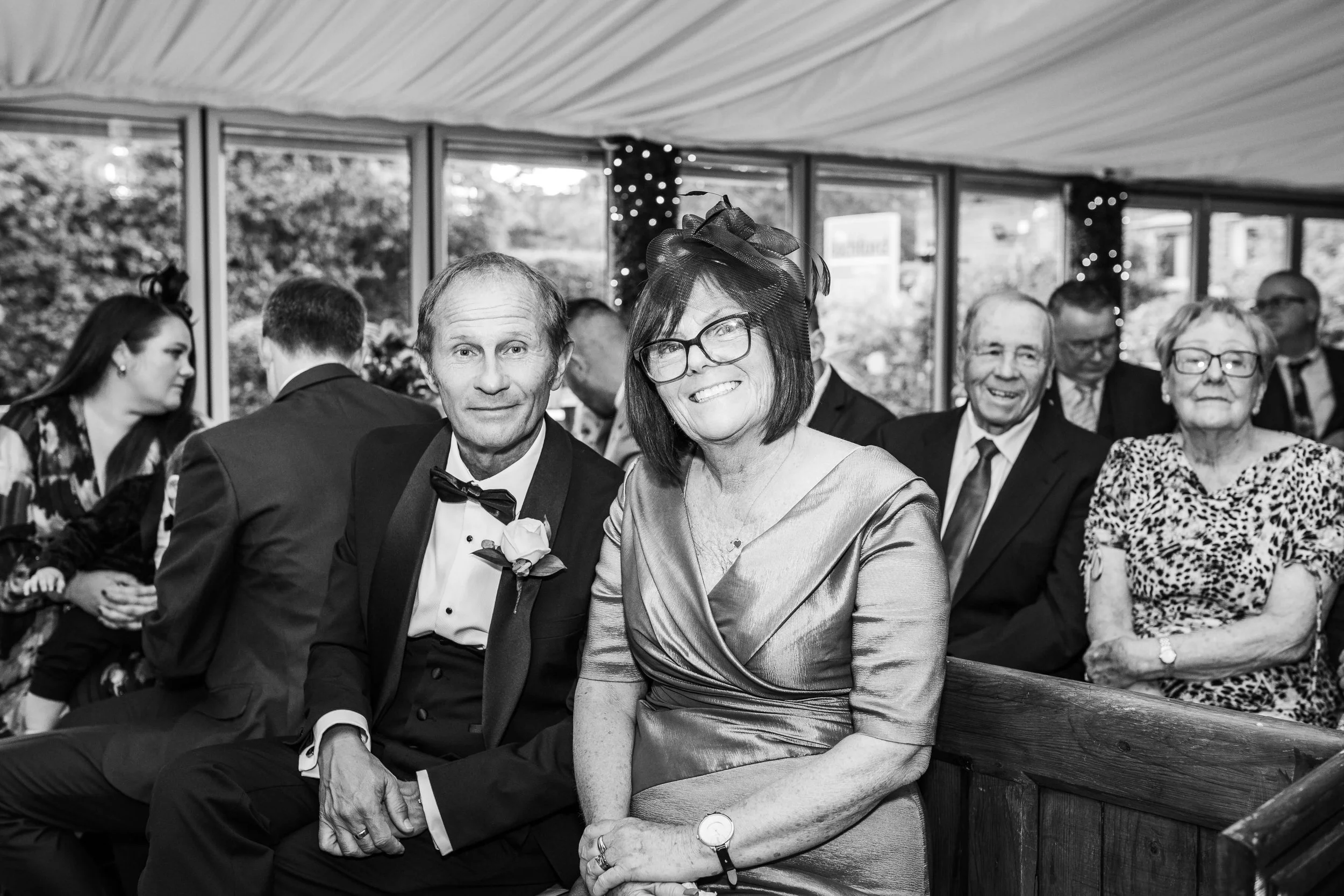 A black and white photo of a group of people dressed in formal attire, seated indoors during a celebration or event. The focus is on a smiling woman with glasses and a hat, seated next to a man in a tuxedo.