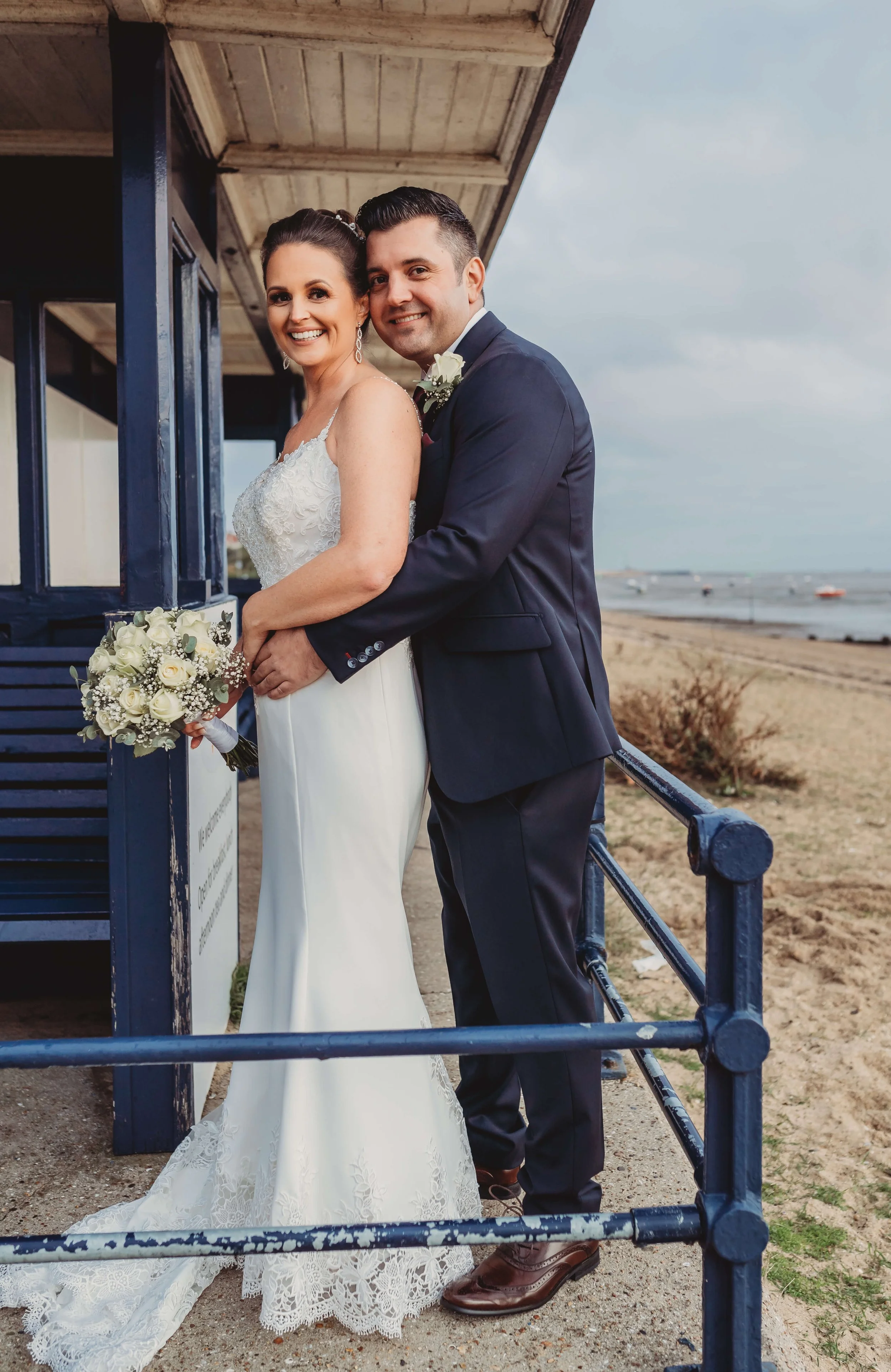Bride and groom standing on a beach near a blue building, smiling at the camera, dressed in wedding attire, holding a bouquet of white roses and greenery.