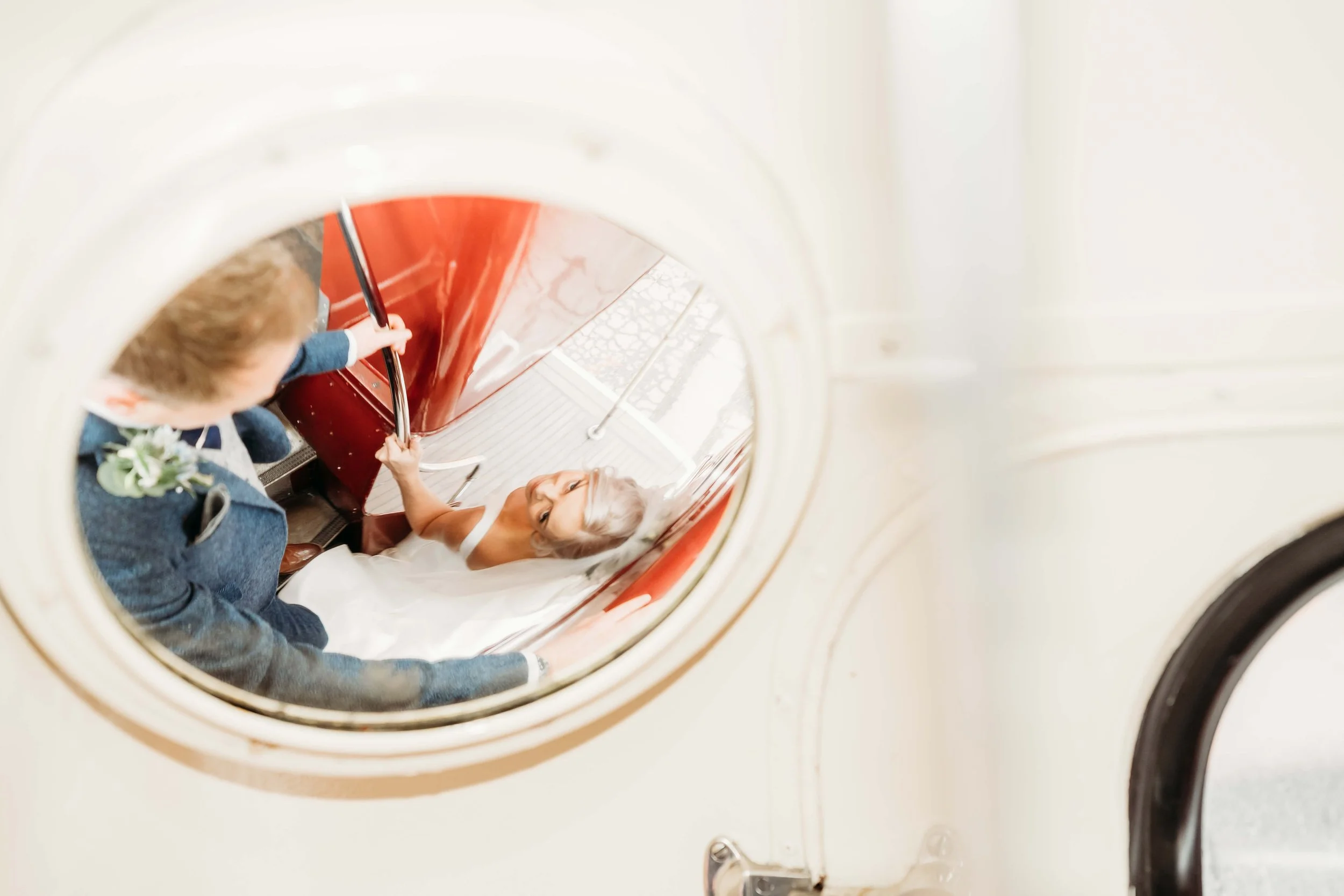 A bride with blonde hair in a white wedding dress lying on a red vintage tram, holding hands with a groom in a blue suit with a boutonniere, as seen from the top of a washer or dryer door.