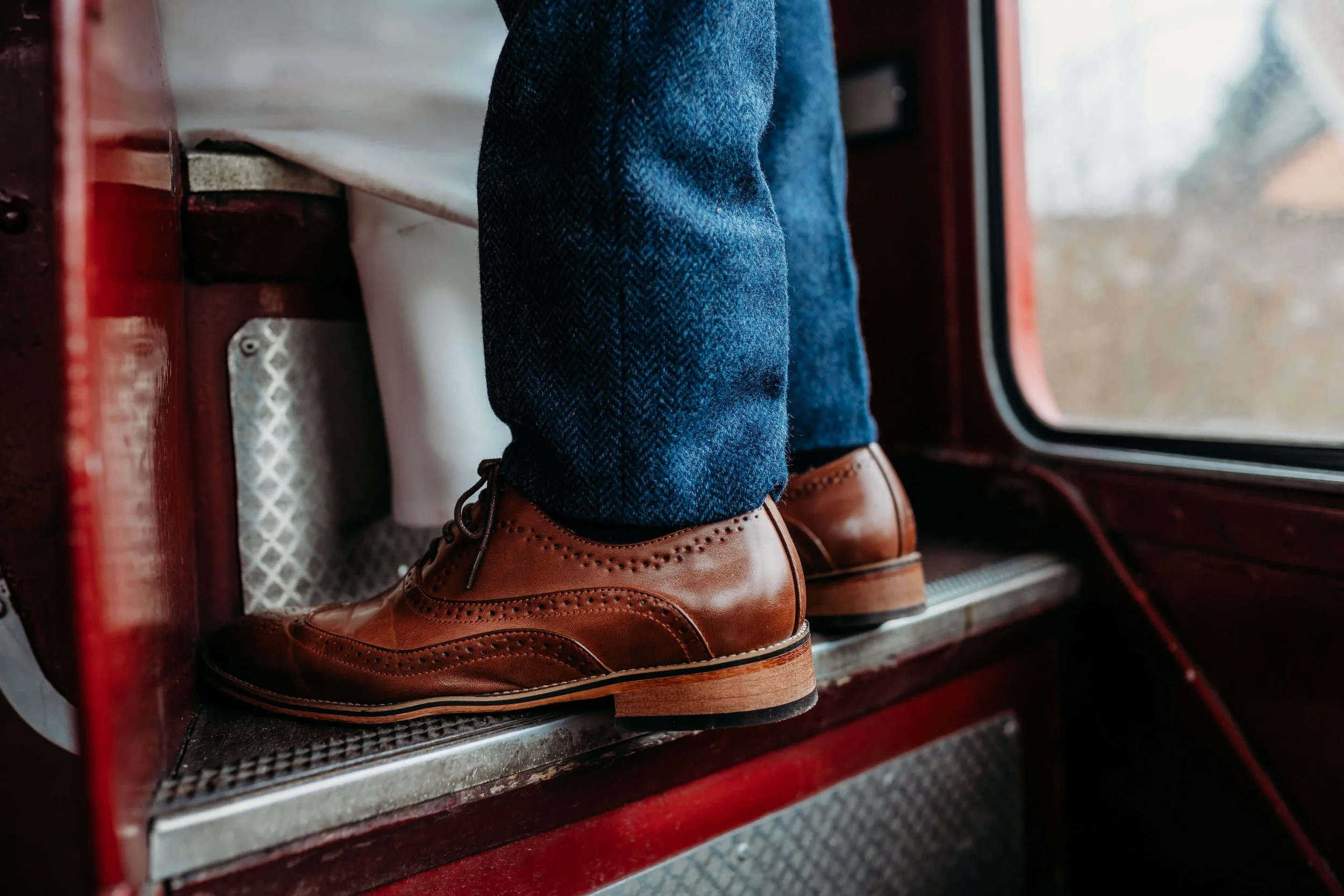 Person wearing brown leather dress shoes and blue jeans standing on the metal step of a vintage red train, with a view of outdoors through a window.