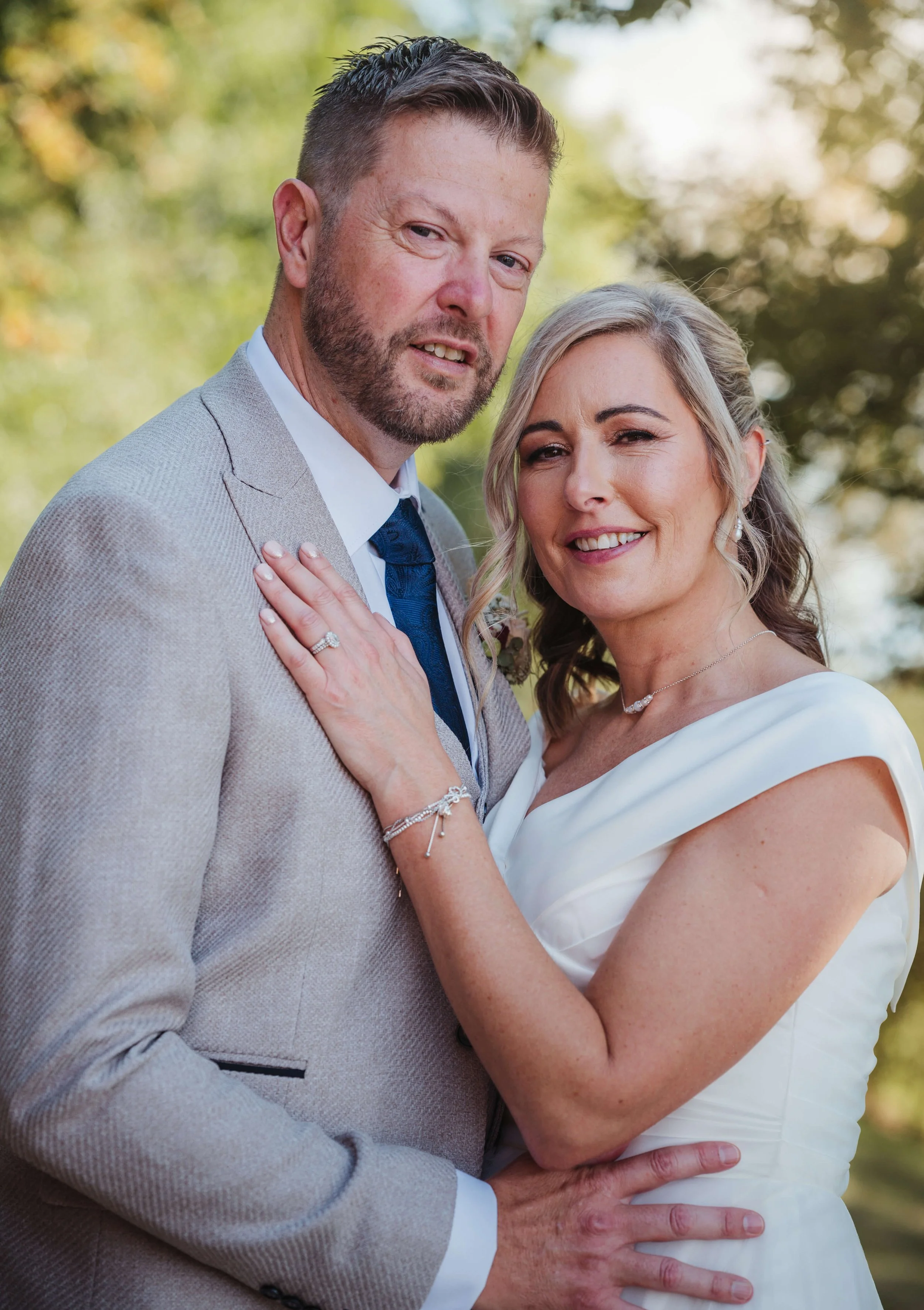 A newlywed couple dressed in wedding attire, standing outdoors with trees in the background. The bride is smiling and wearing a white dress, jewelry, and has her hand on the groom's chest. The groom is in a light beige suit, white shirt, and dark tie