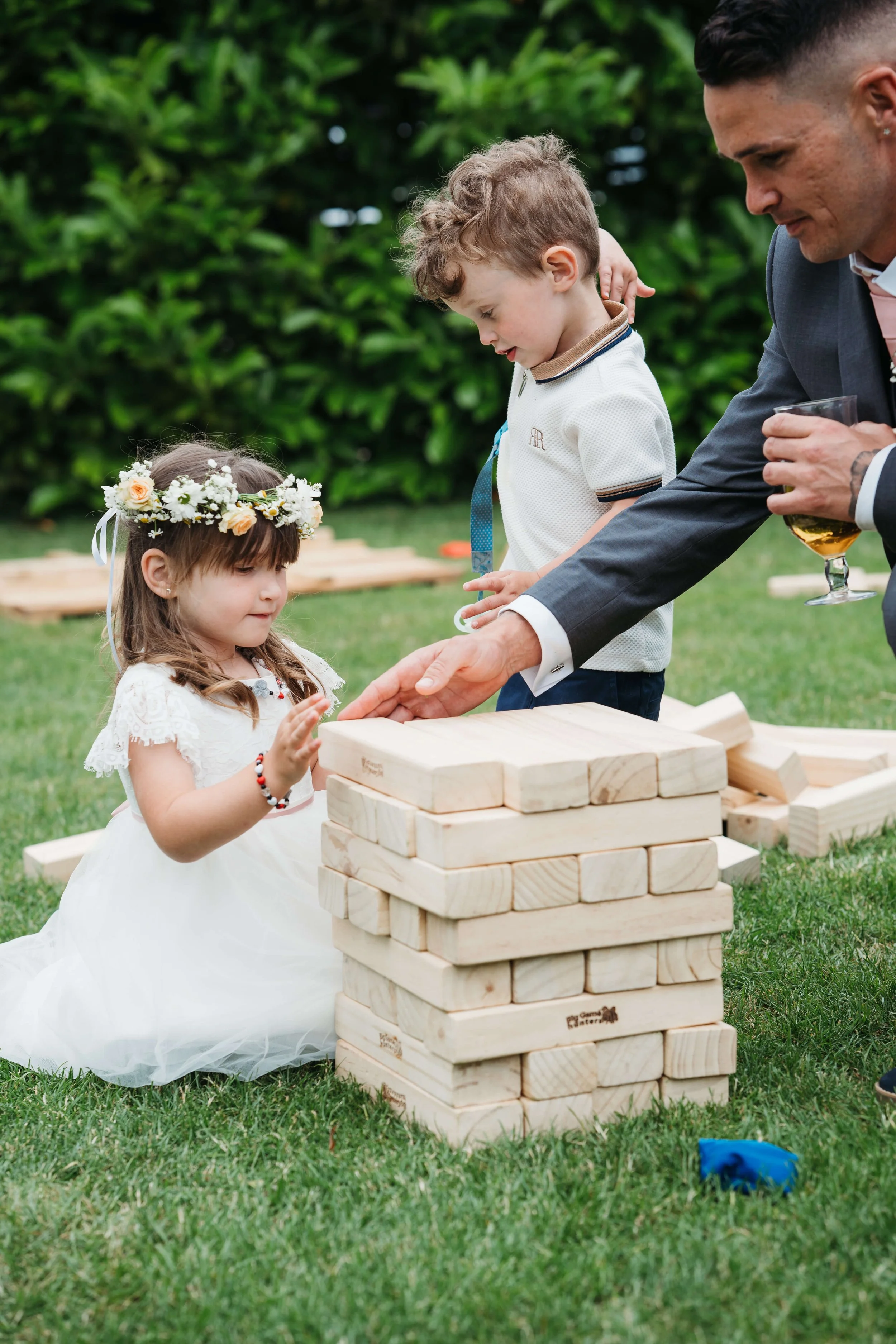 A young girl in a white dress with a floral crown is playing a giant Jenga game with two boys and an adult male. She is sitting on grass in an outdoor setting with green trees in the background.