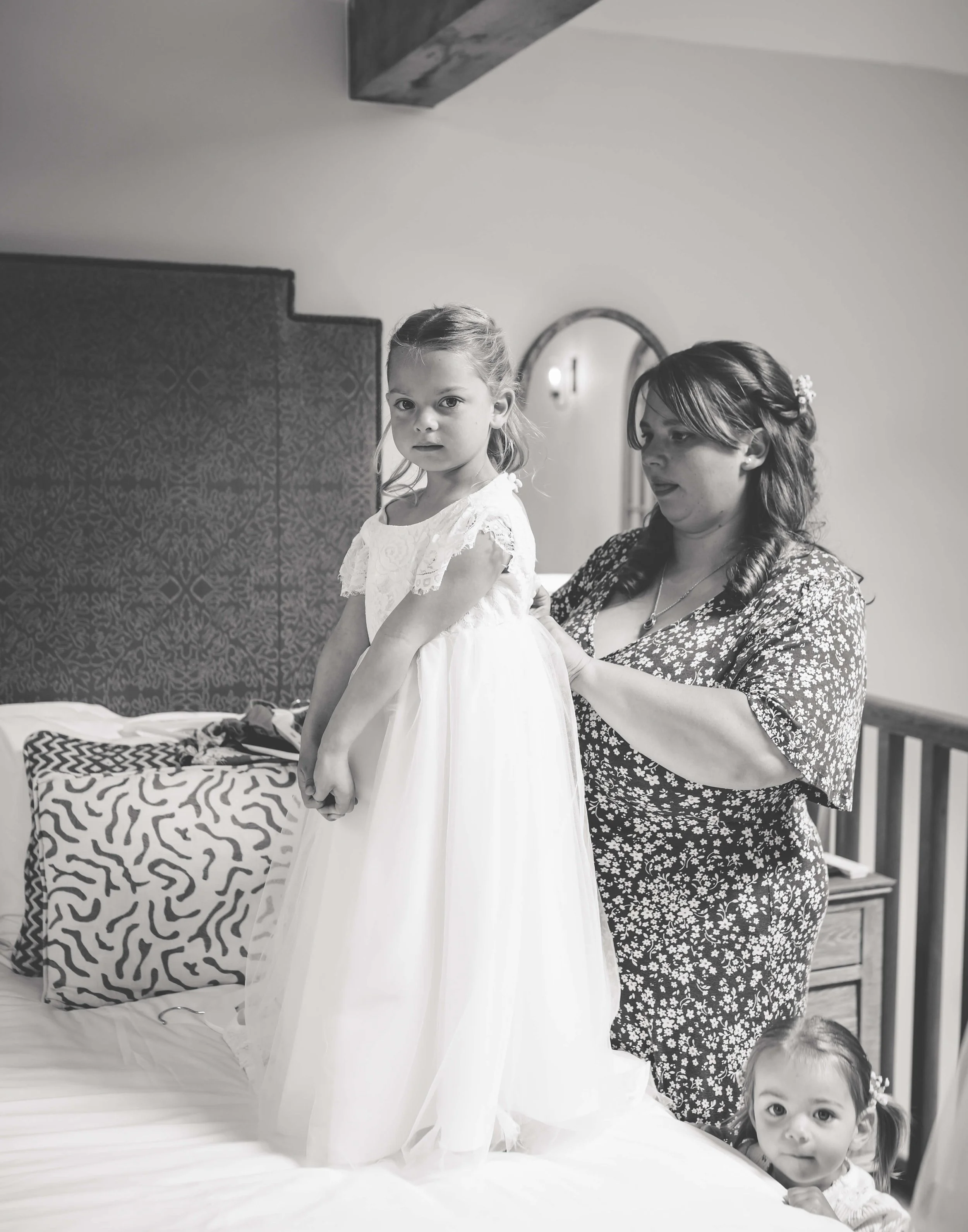 A woman helps a young girl in a white dress stand on a bed, with a smaller girl peeking from behind the bed.