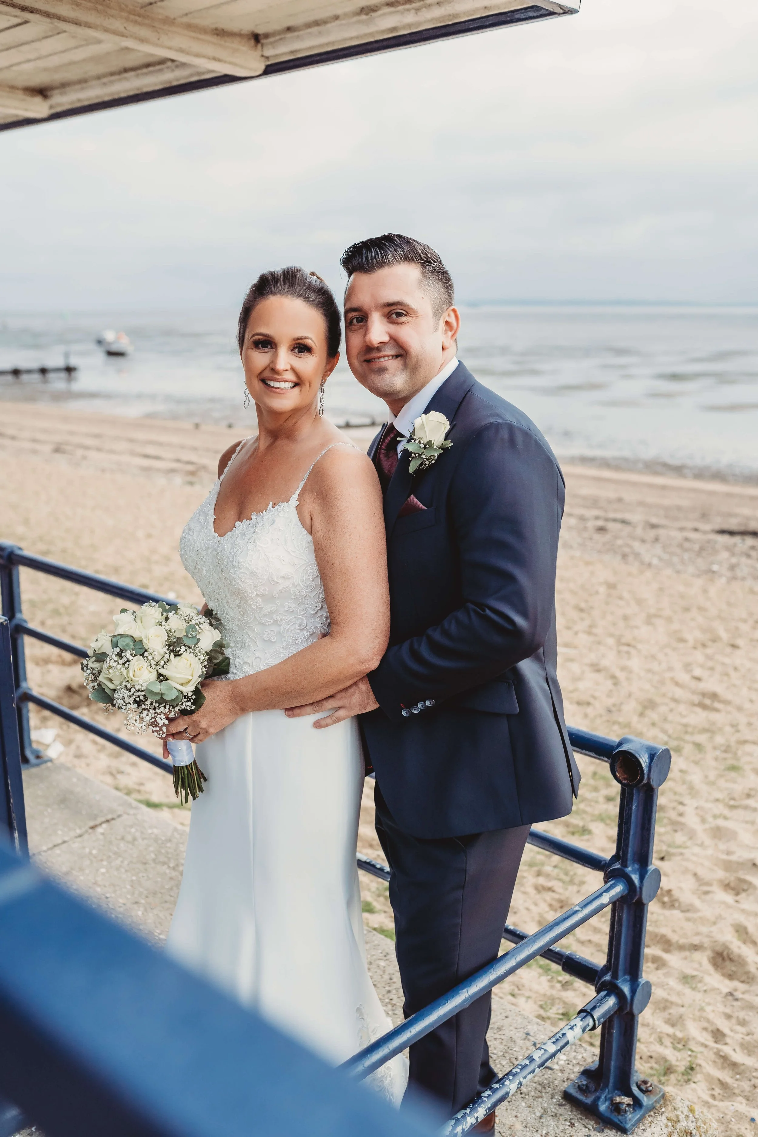 A newlywed couple on the beach, with the bride holding a bouquet of white roses and the groom in a navy suit with a white rose boutonniere, standing under a shaded area near the ocean.