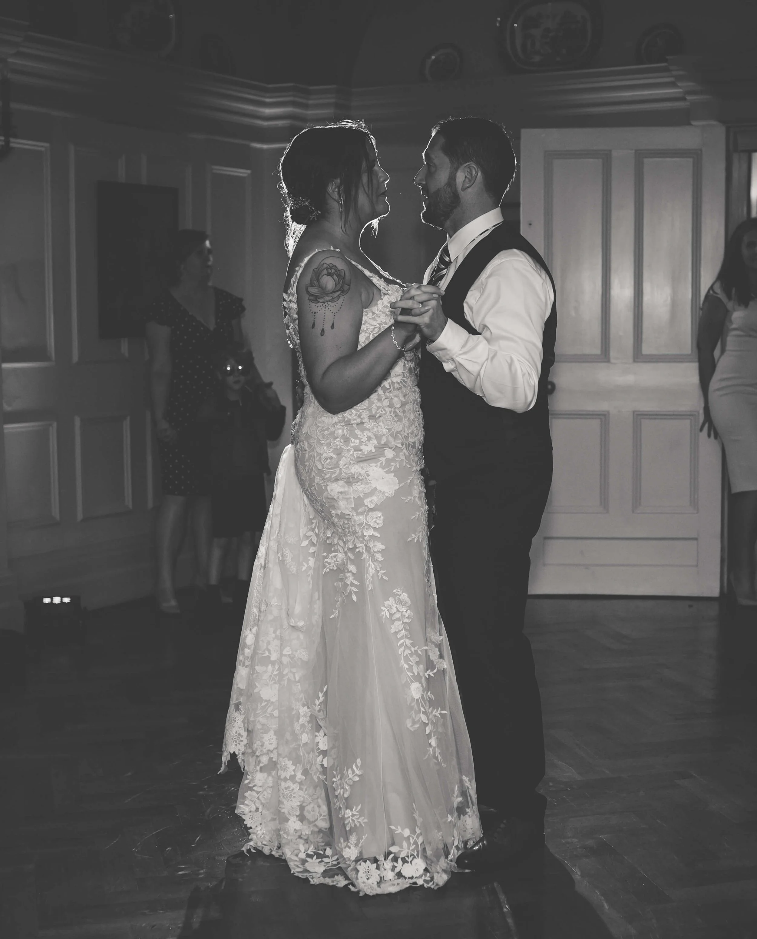 A bride and groom are dancing closely during their wedding reception, with onlookers watching in the background, black and white photo.