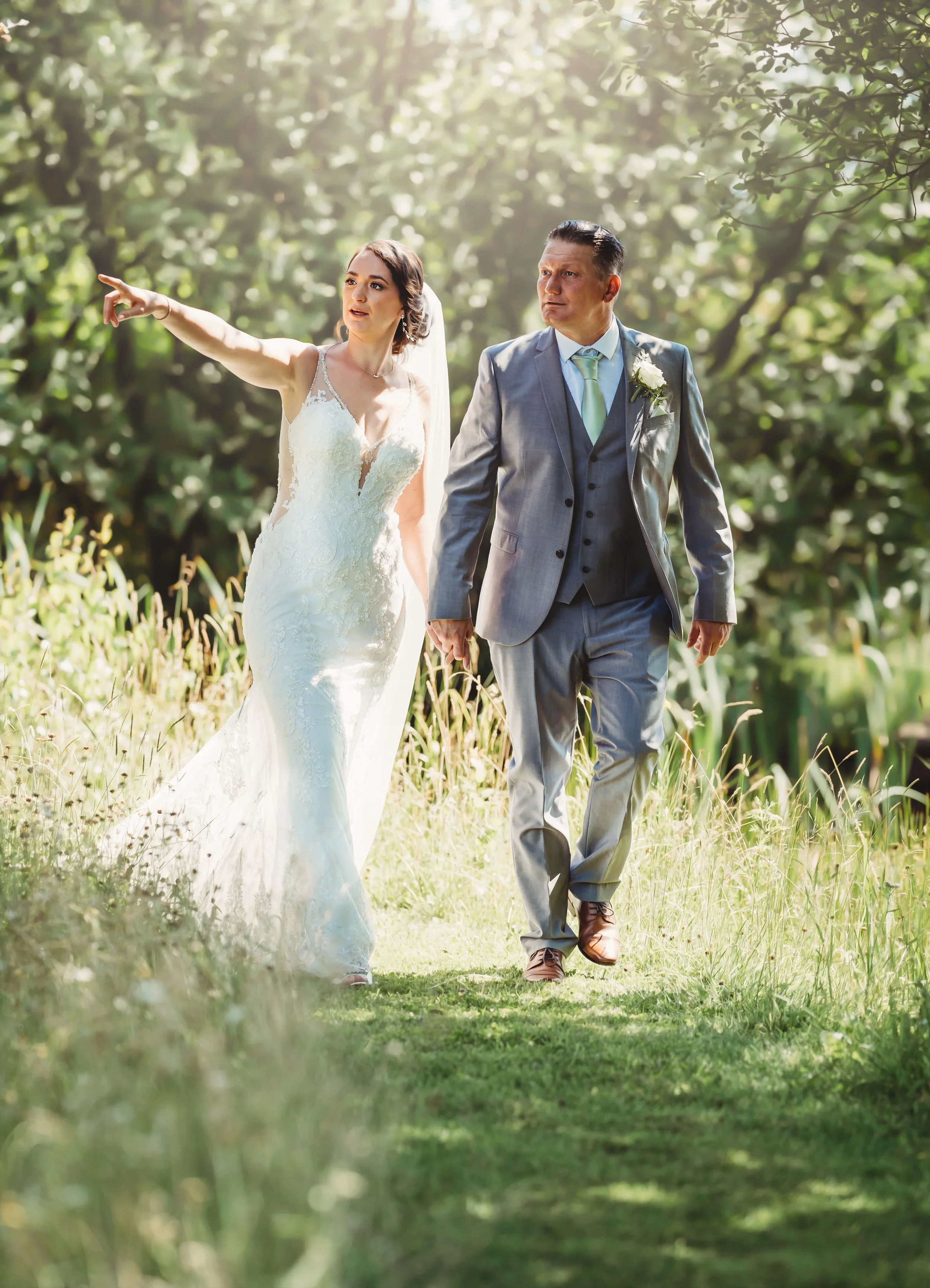A bride and an older man, likely her father, walking through a lush, green outdoor setting on her wedding day. The bride is wearing a white lace wedding gown and pointing, while the man is dressed in a gray suit with a light blue tie, holding her han