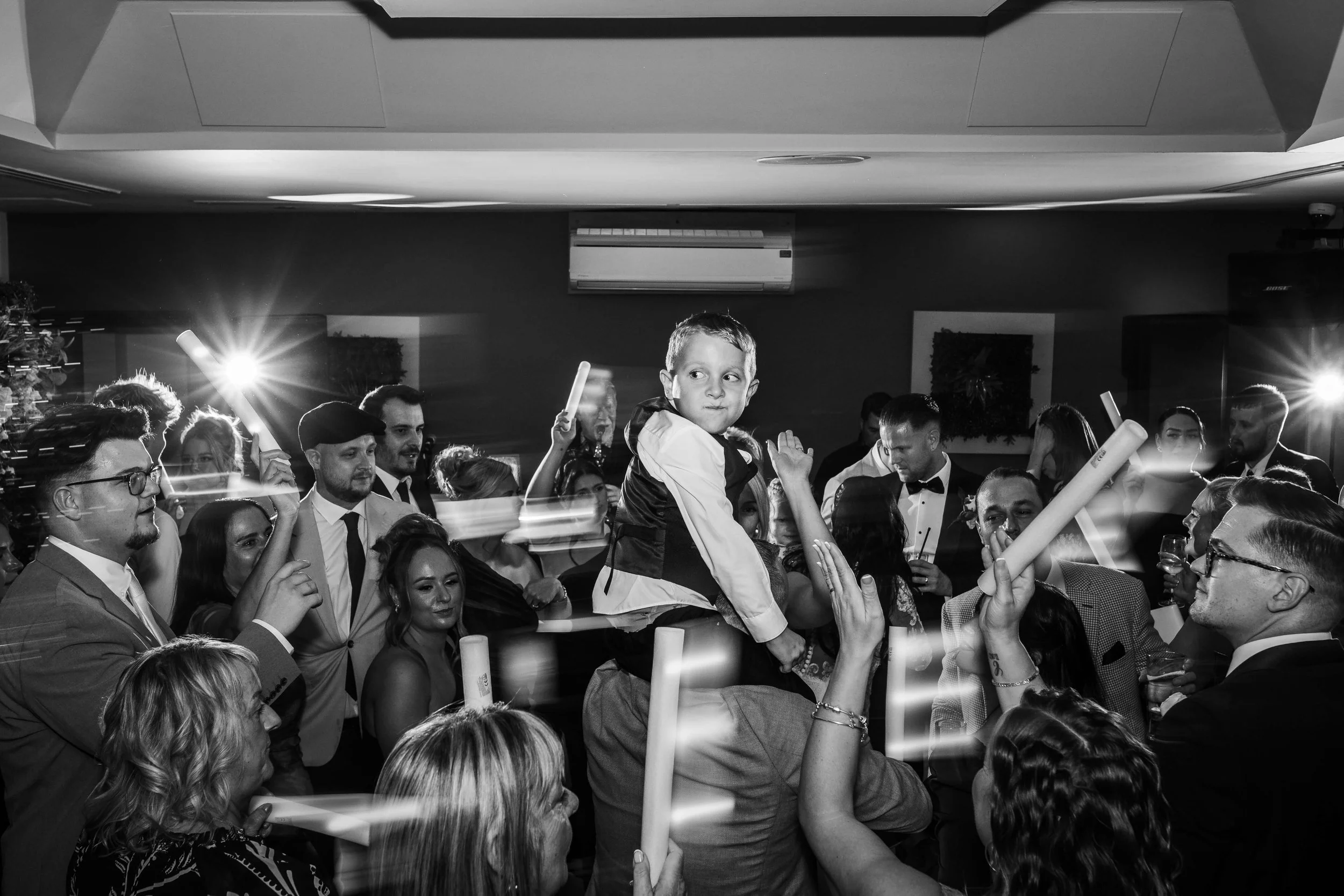 A black-and-white photo of a party with many people, including a young boy sitting on someone's shoulders, surrounded by adults holding glow sticks, some taking photos, in an indoor venue.