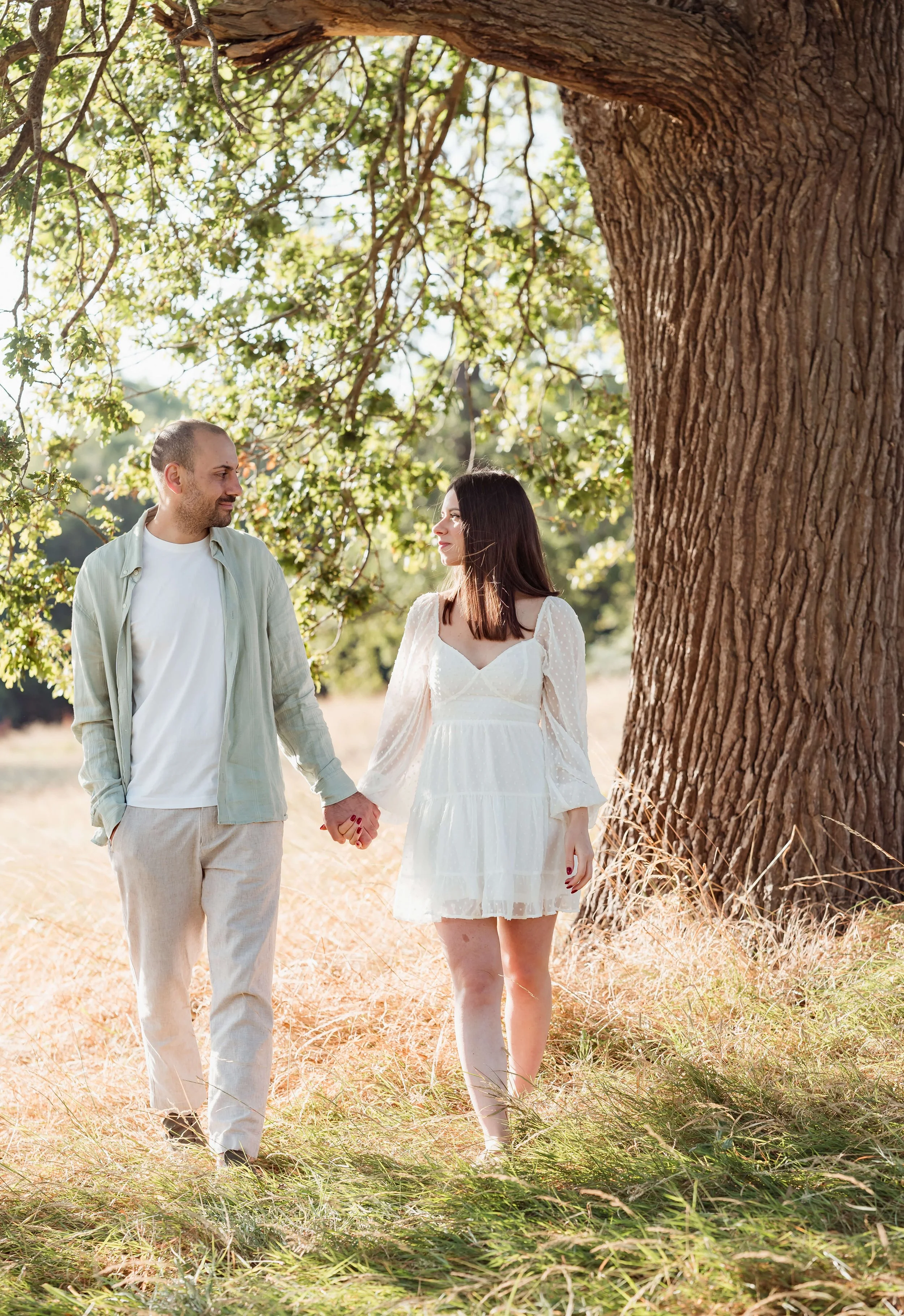 A man and woman holding hands while walking outdoors in a grassy field near a large tree with green leaves. The man is wearing a light-colored shirt and pants, and the woman is wearing a white dress. They are looking at each other.