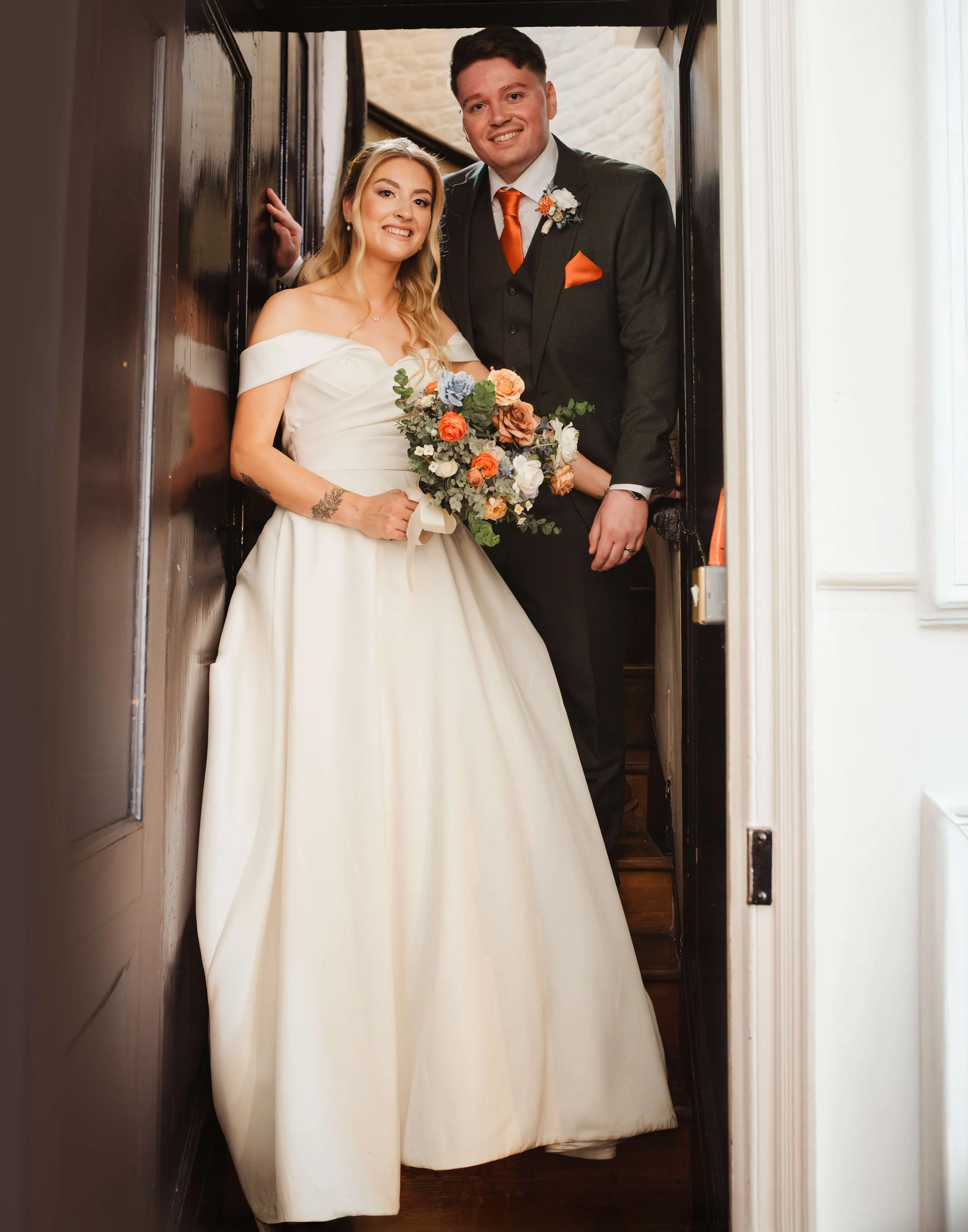 A bride and groom standing in a doorframe, smiling for a wedding photo. The bride is wearing an off-shoulder white wedding gown and holding a bouquet of colorful flowers. The groom is wearing a black suit with an orange tie and matching pocket square