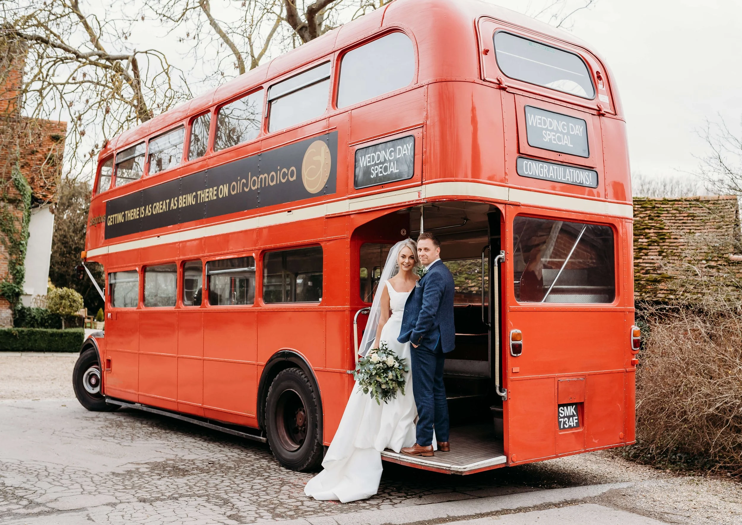 A bride and groom standing together in front of a red double-decker bus decorated for a wedding, with wedding-related signs displayed on the bus.