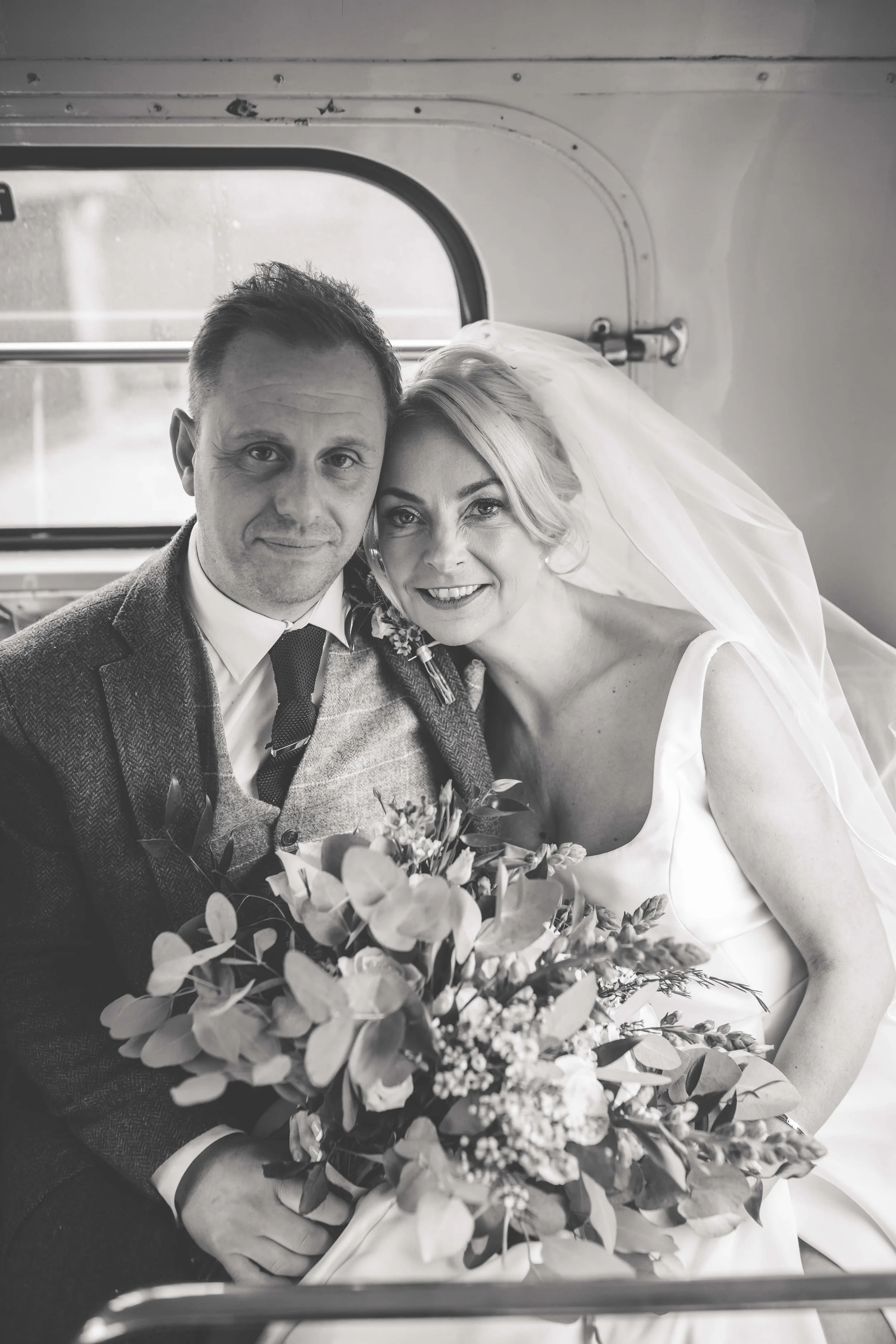 Black and white photo of a bride and groom sitting inside a vehicle, smiling at the camera. The bride is wearing a wedding dress and veil, holding a bouquet. The groom is in a suit with a tie.