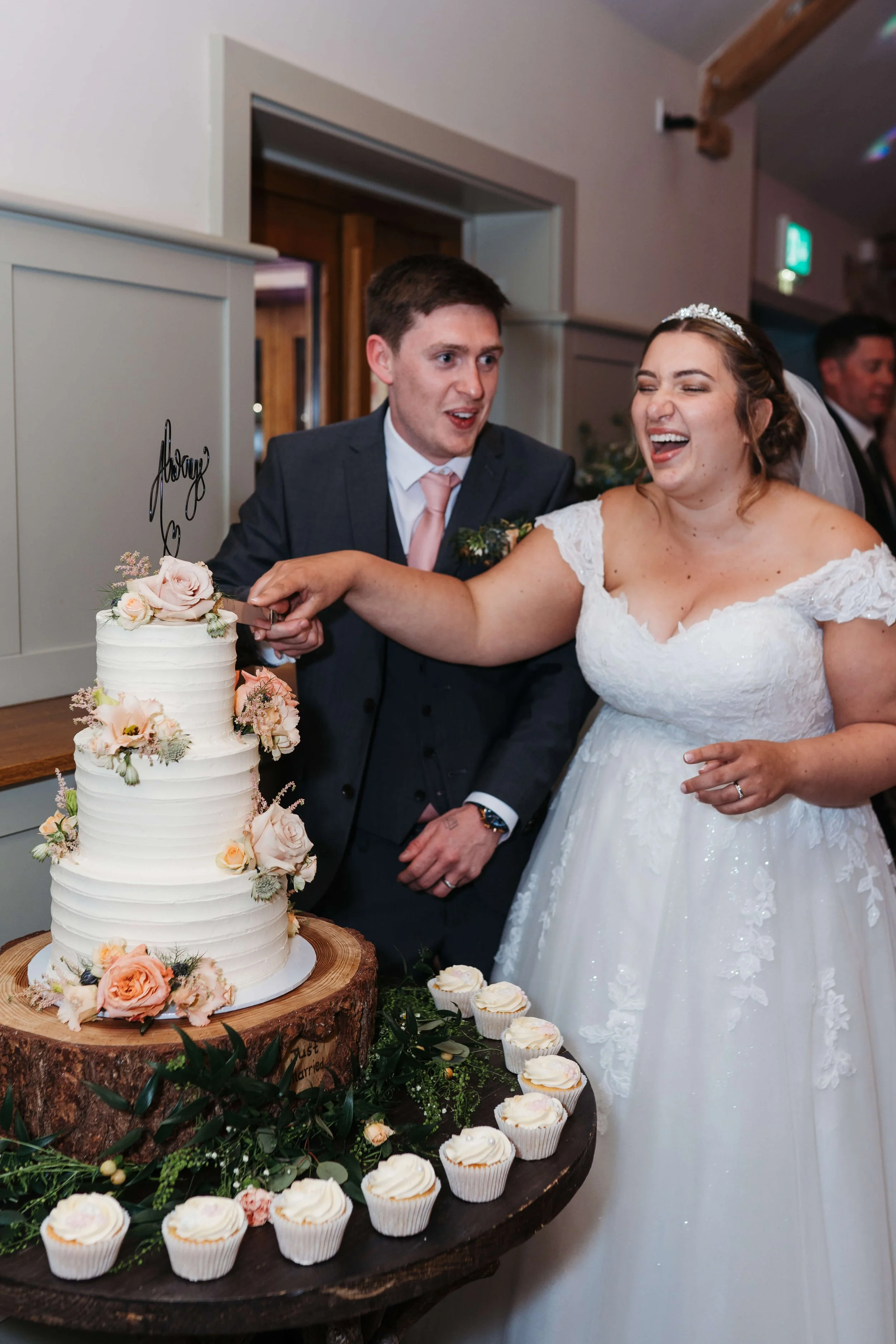 Bride and groom cutting a wedding cake. The cake is decorated with flowers and set on a wooden table with cupcakes around it.