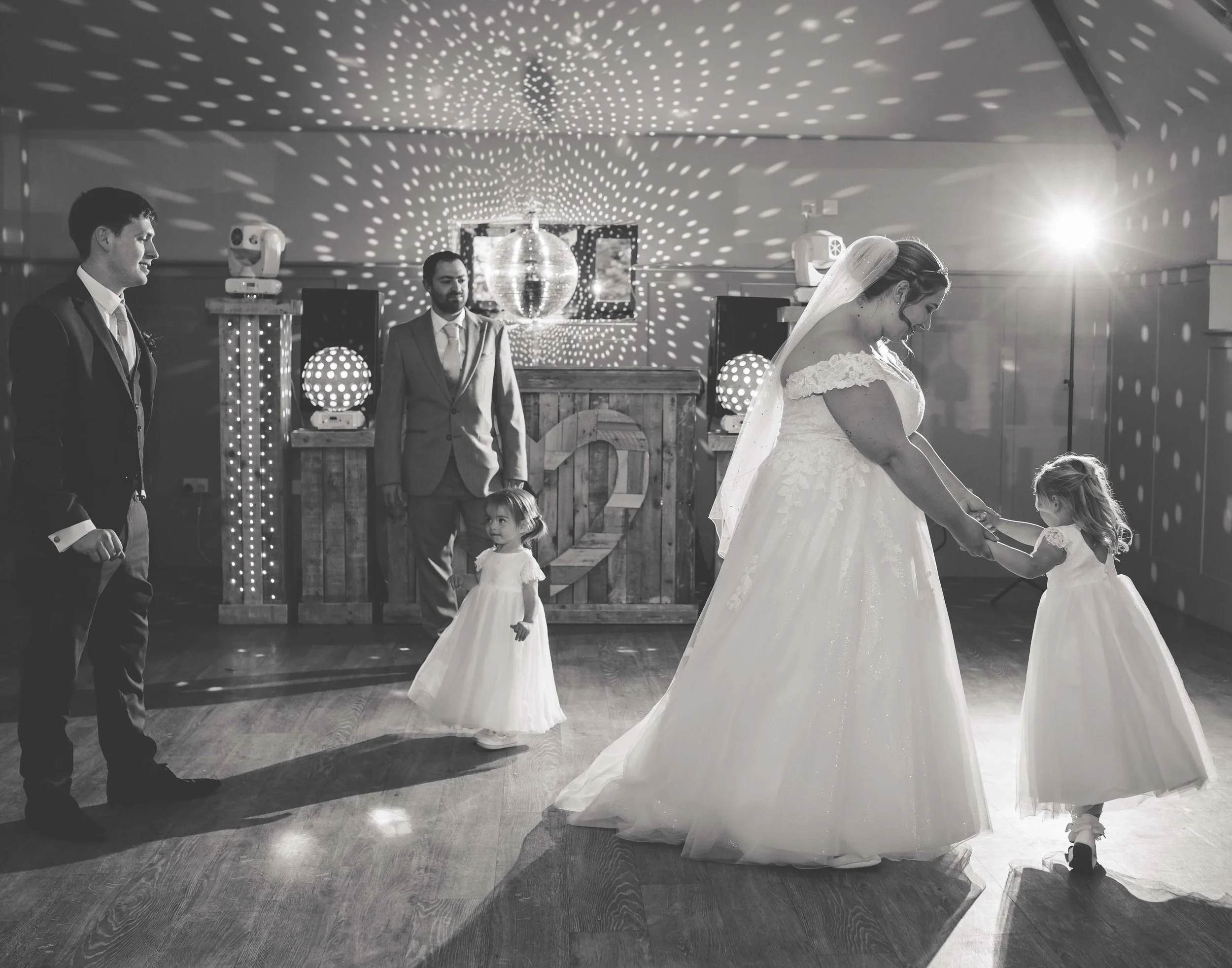 A black and white photo of a wedding reception dance floor with a bride in a gown holding hands with a small girl in a dress. Two young girls in white dresses and two men in suits are observing. A disco ball and decorative lights are in the backgroun