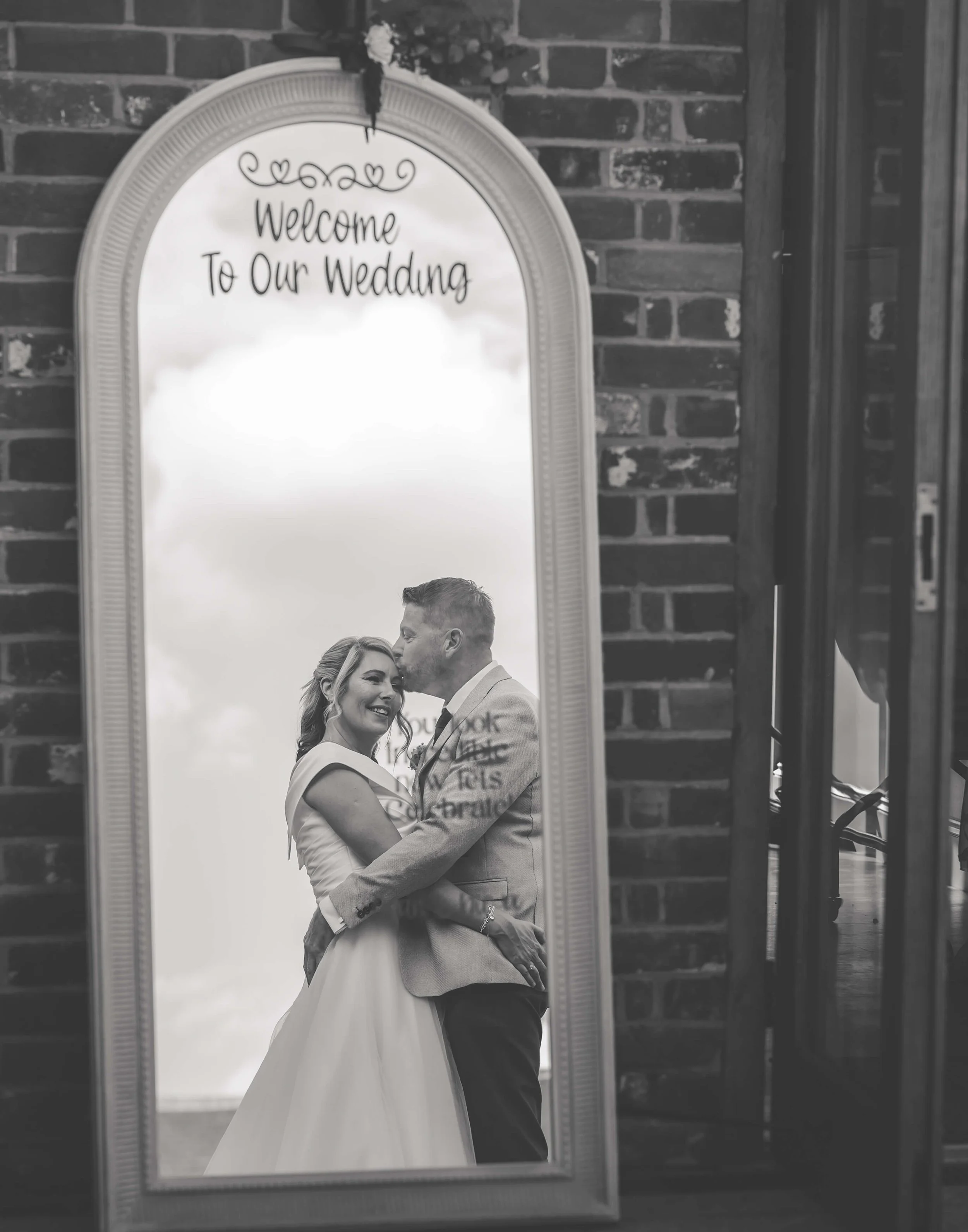 A wedding couple smiling and embracing their reflection in a mirror, with a sign at the top that reads 'Welcome To Our Wedding' and a brick wall in the background.