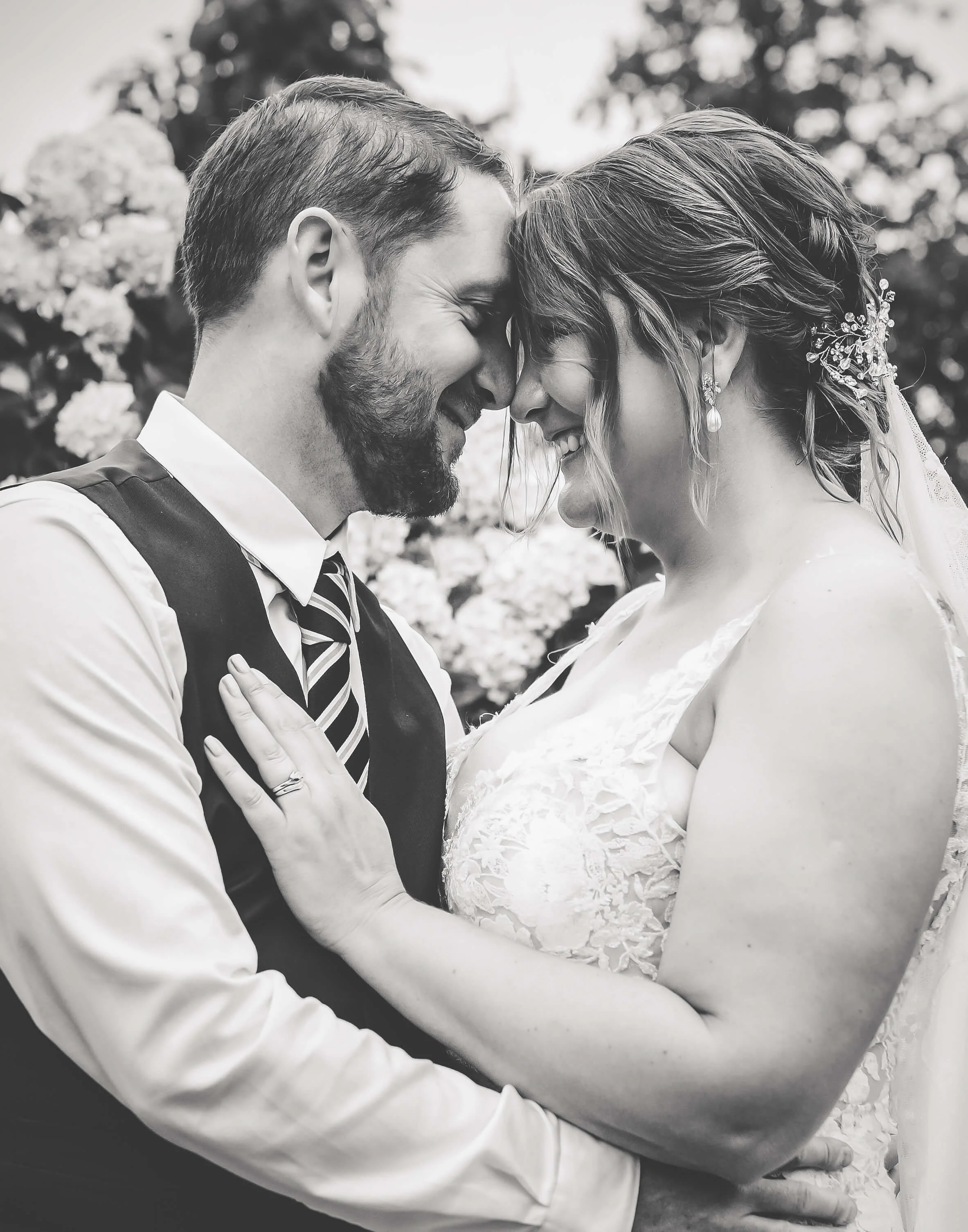 A black and white photo of a smiling bride and groom touching foreheads, holding each other at a wedding.