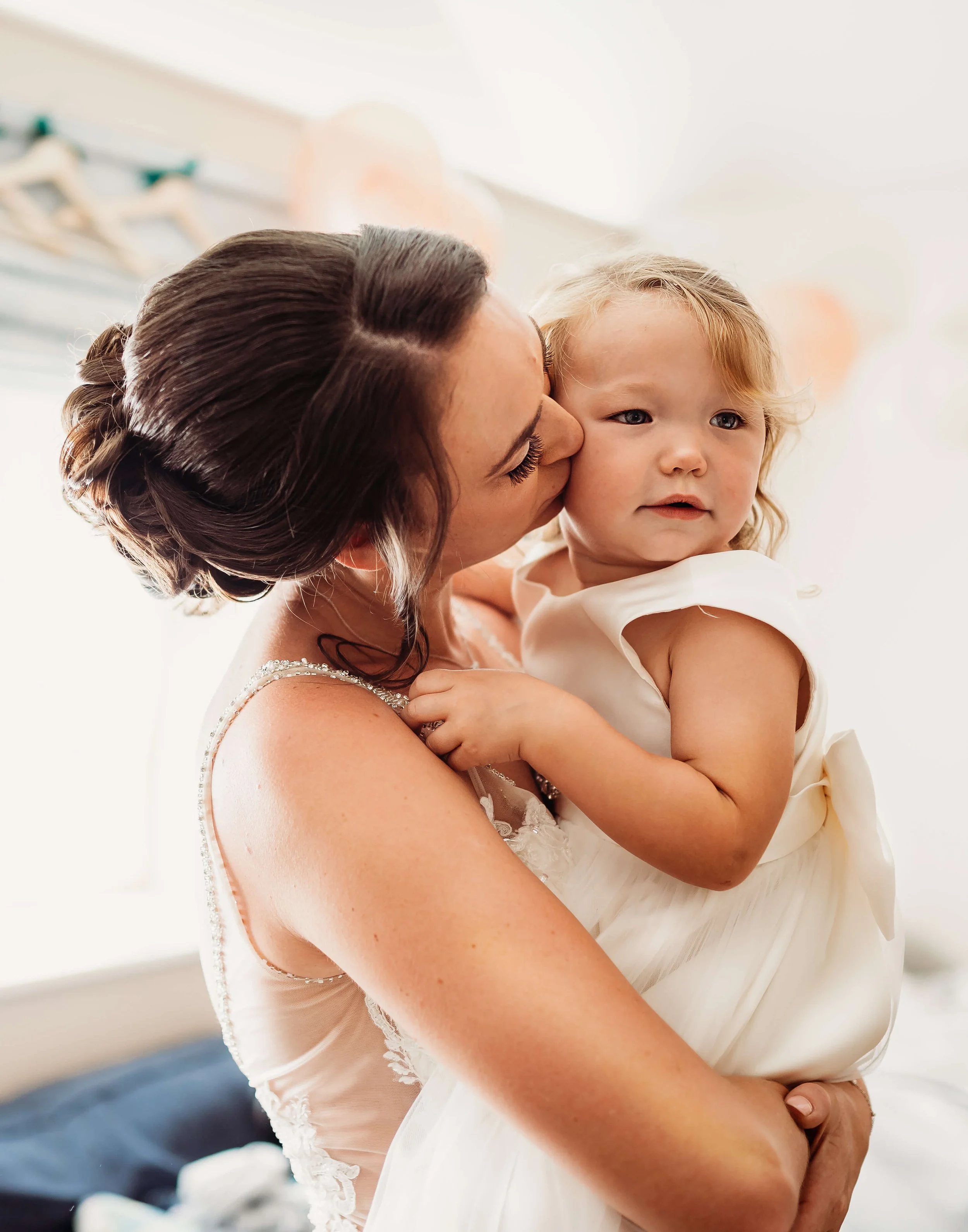 A woman in a wedding dress holding and kissing a young girl in a white dress.