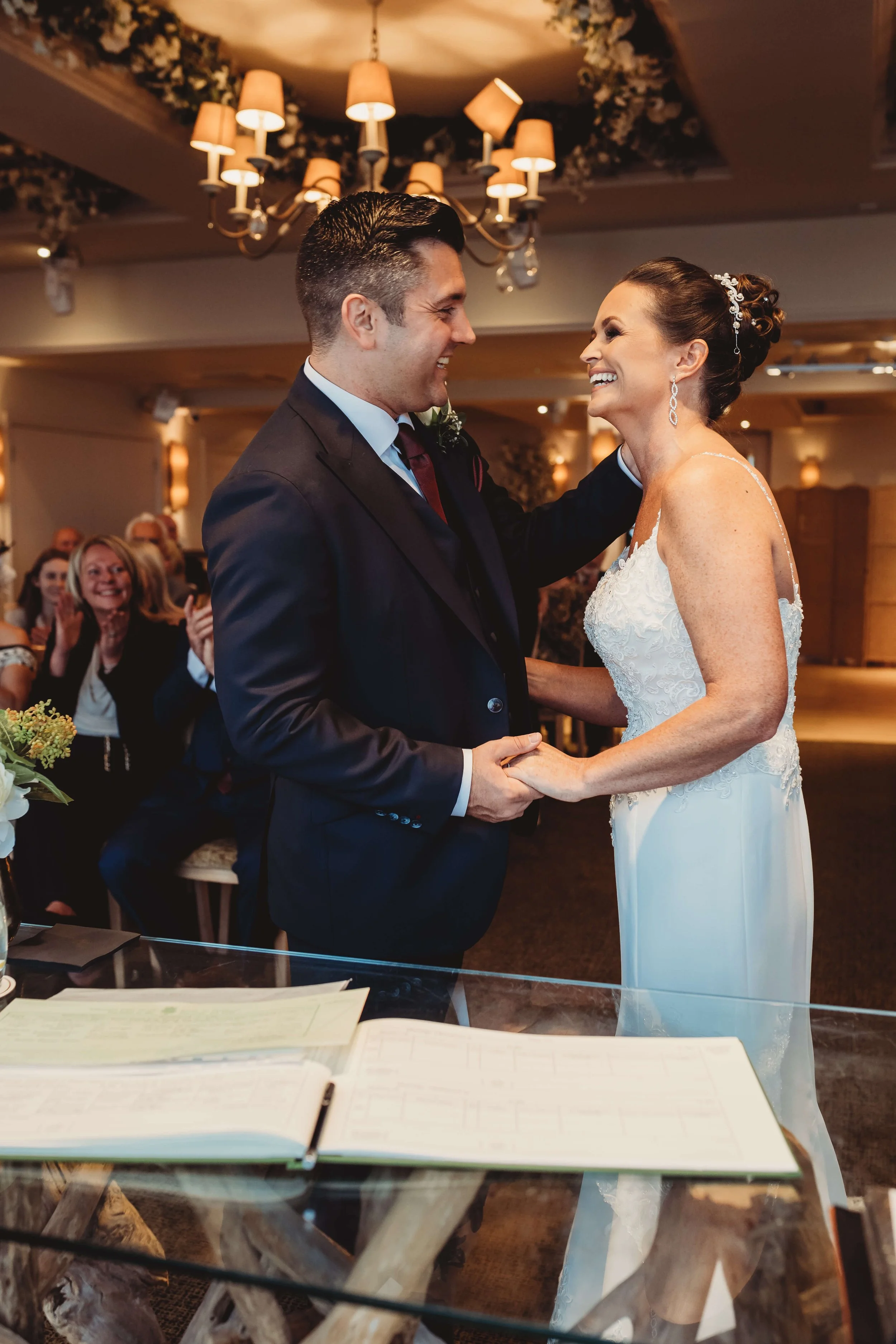 A bride and groom holding hands and smiling during their wedding ceremony, with guests clapping in the background.