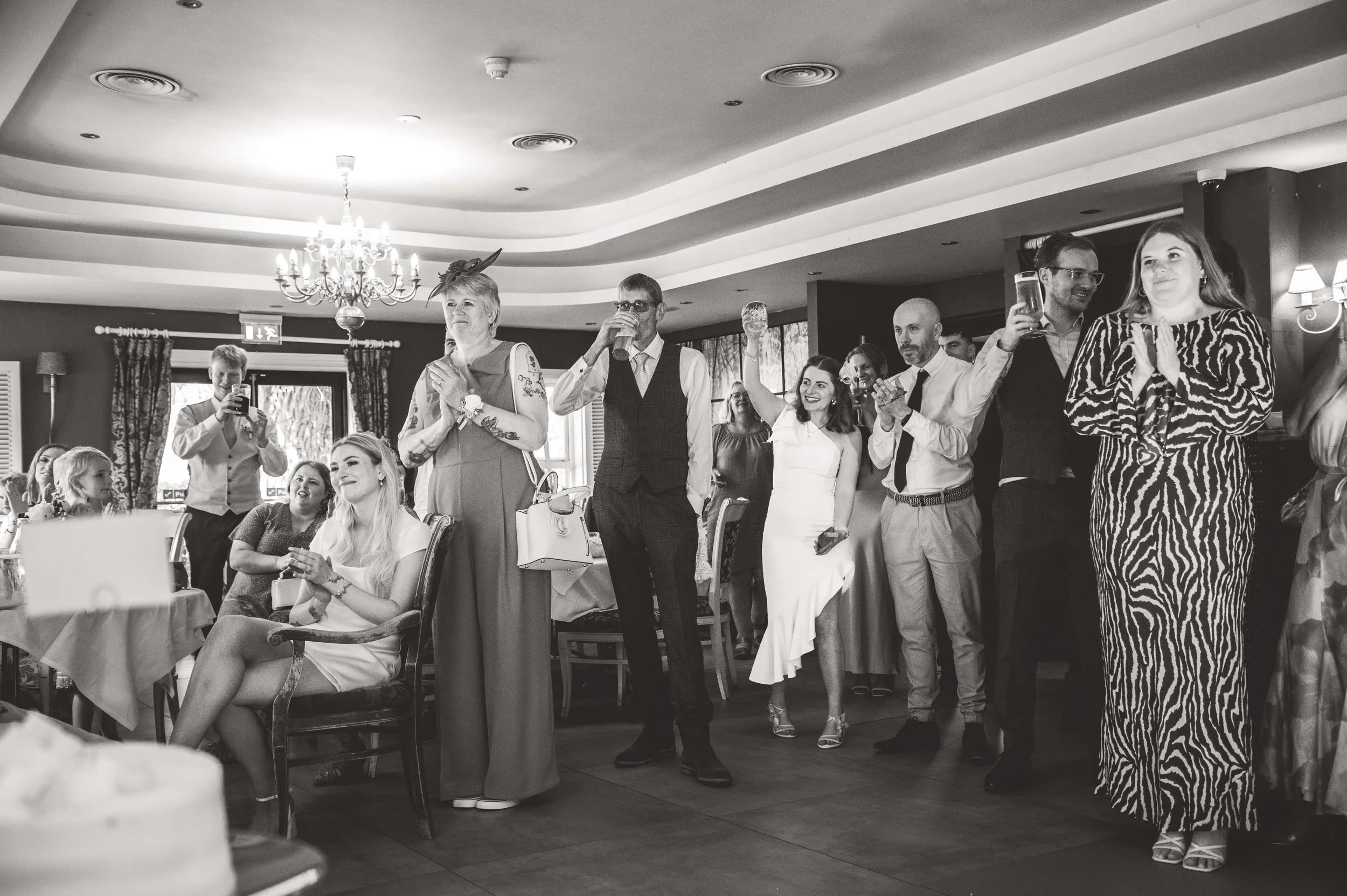 People at a celebration party, some standing and some seated, clapping and holding drinks, with a decorated ceiling and windows in the background.