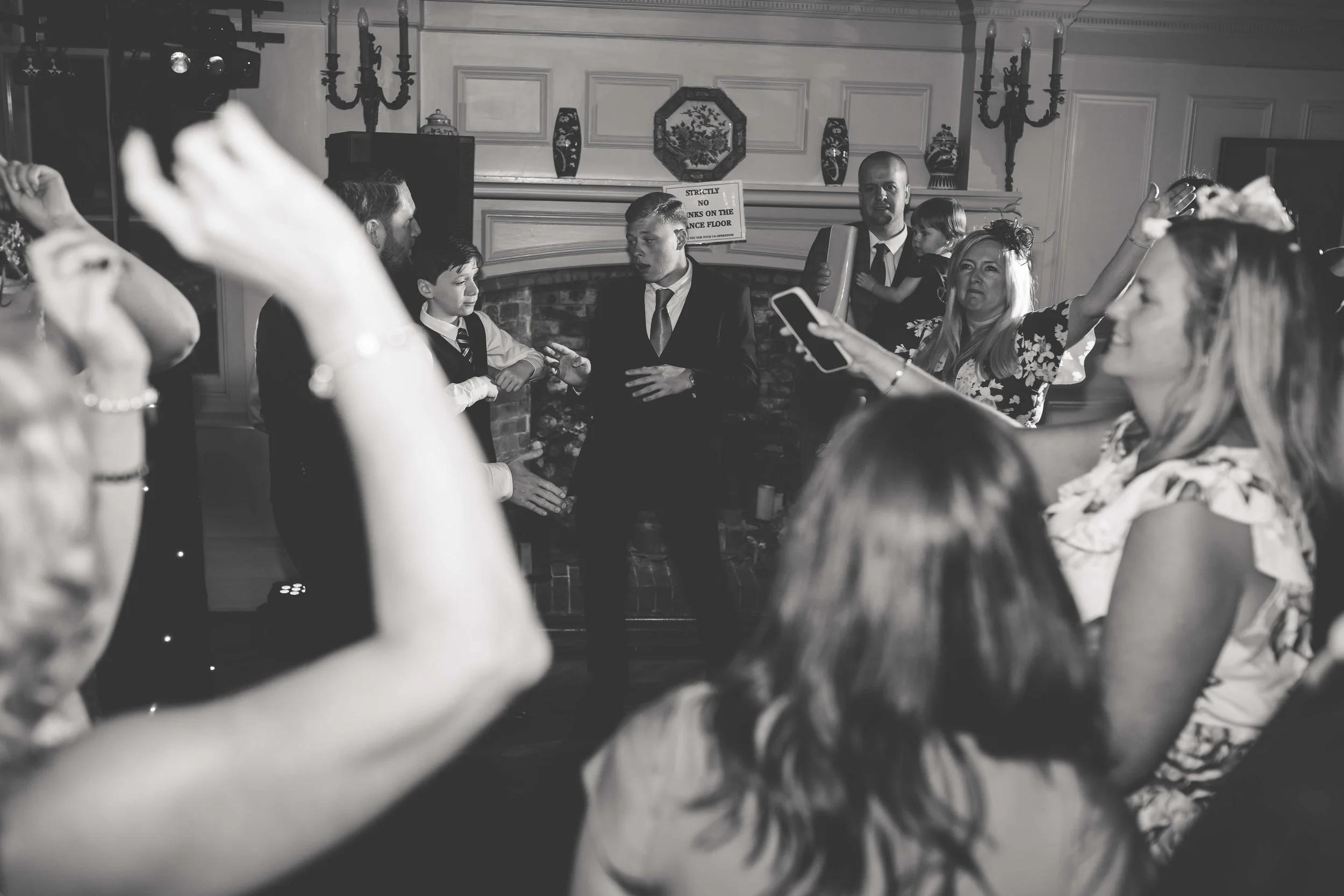 Black and white photo of a group of people dancing and socializing indoors, with a fireplace and decorative wall art in the background.