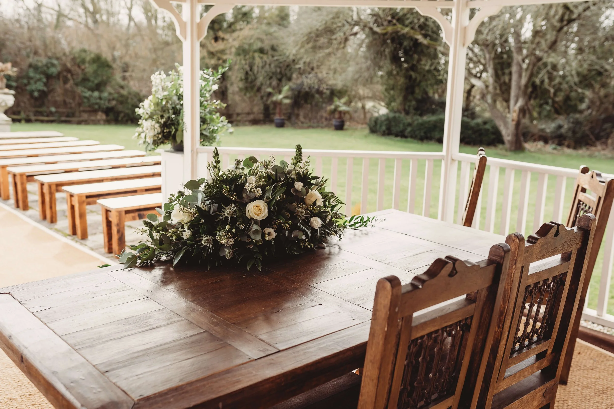Wooden outdoor dining table with a floral centerpiece, set on a porch with white railing, overlooking a grassy yard with trees.