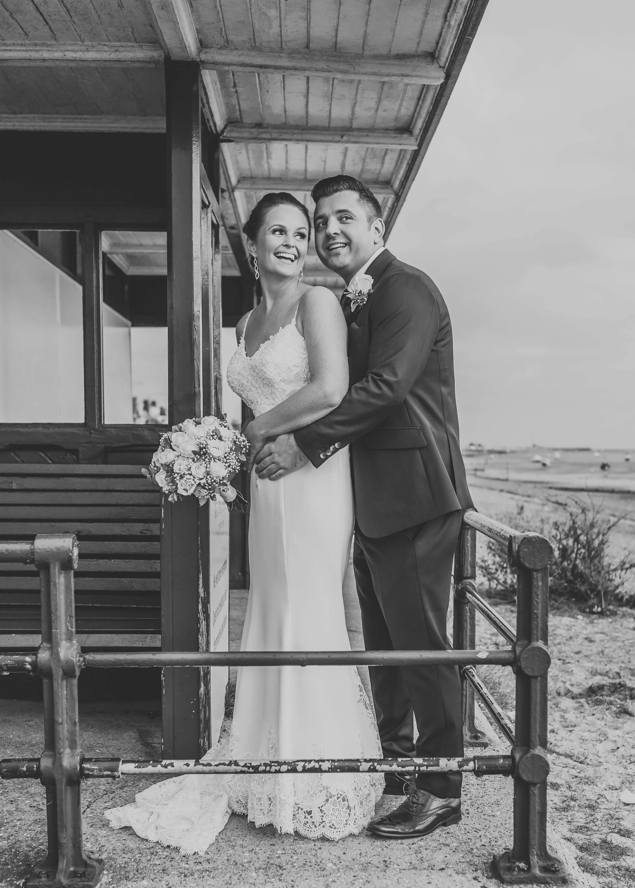 Black and white photo of a bride and groom standing outside on a beach, smiling and embracing each other, with the ocean in the background.
