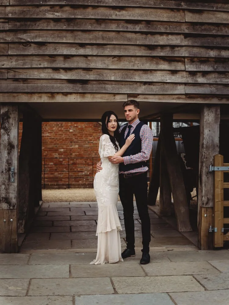A man and woman, dressed in formal attire, standing close together outside a rustic wooden structure with a brick wall in the background.