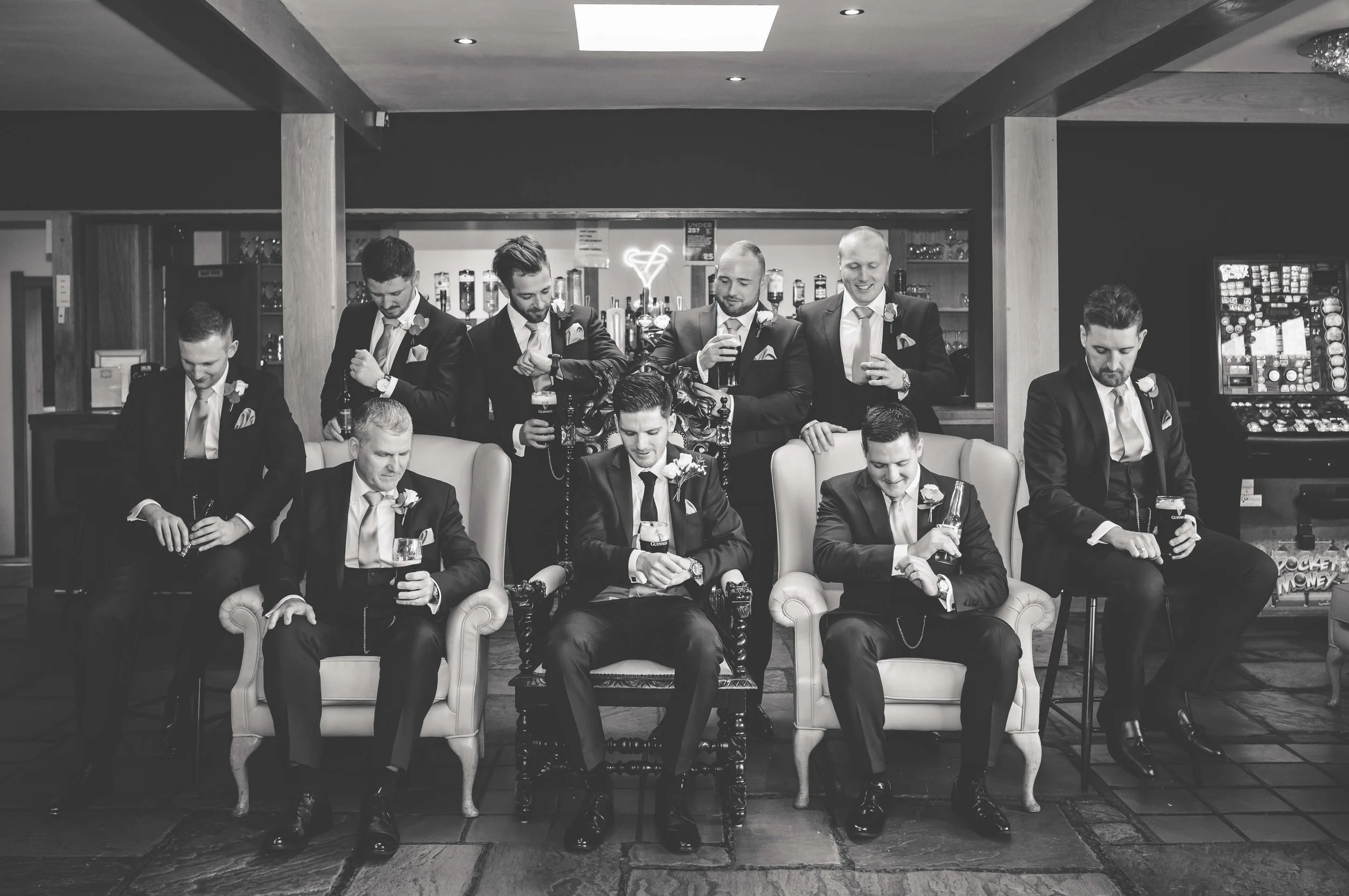 Black and white photo of ten men dressed in suits, some sitting and some standing, in a bar or lounge area, holding drinks and posing.