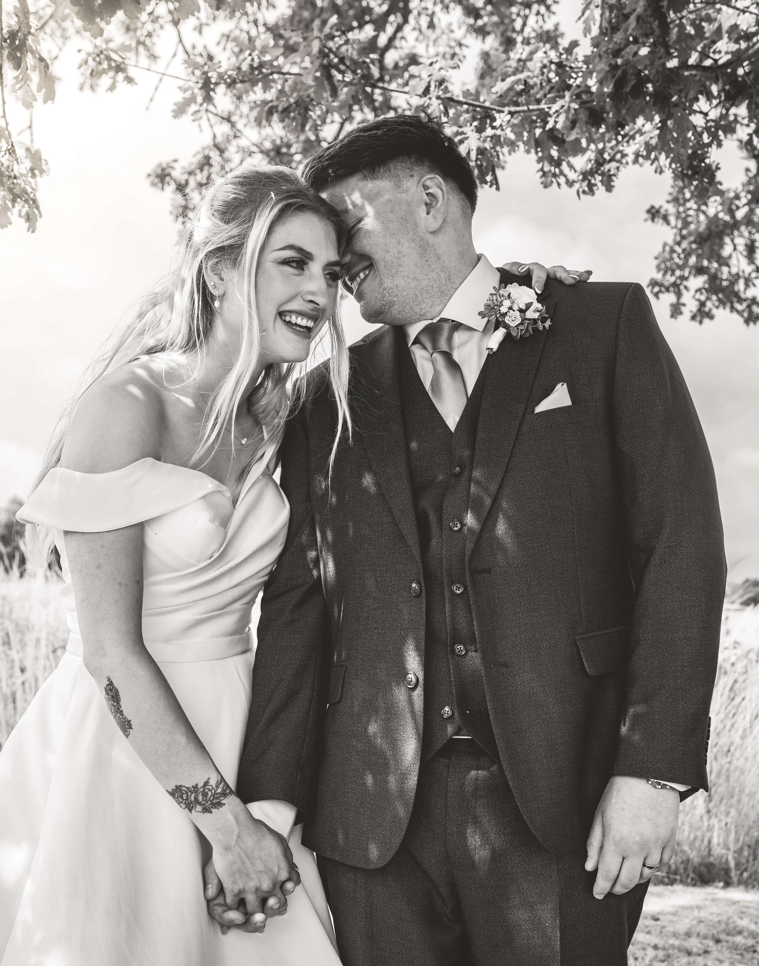Black and white photo of a happy couple in wedding attire holding hands, leaning their foreheads together and smiling outdoors.