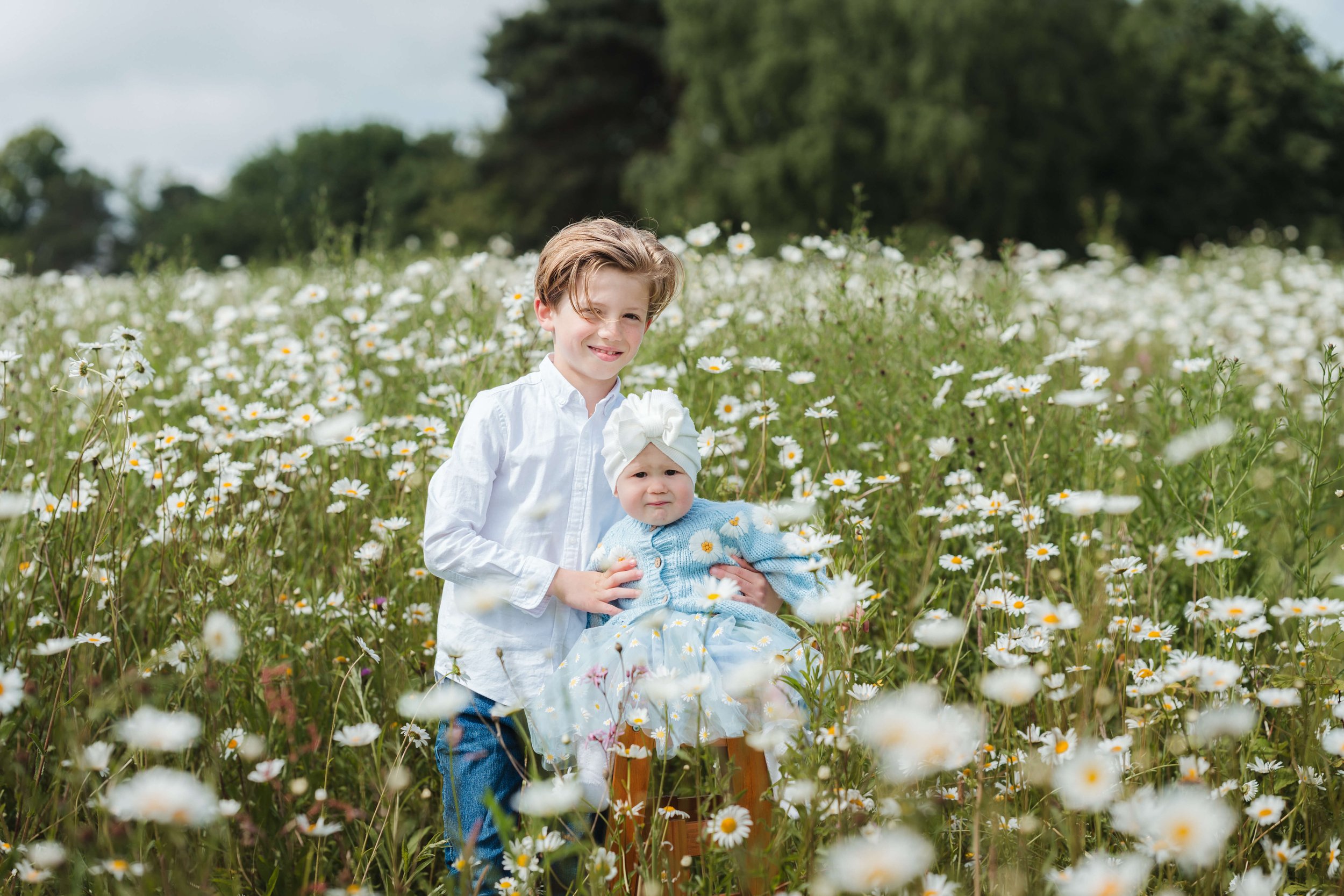 Two young children, a boy and a baby girl, in a field of white daisies. The boy is smiling and wearing a white shirt and jeans, holding the baby girl who is dressed in a blue dress, sweater, and a white hat. The background shows trees and a cloudy sk