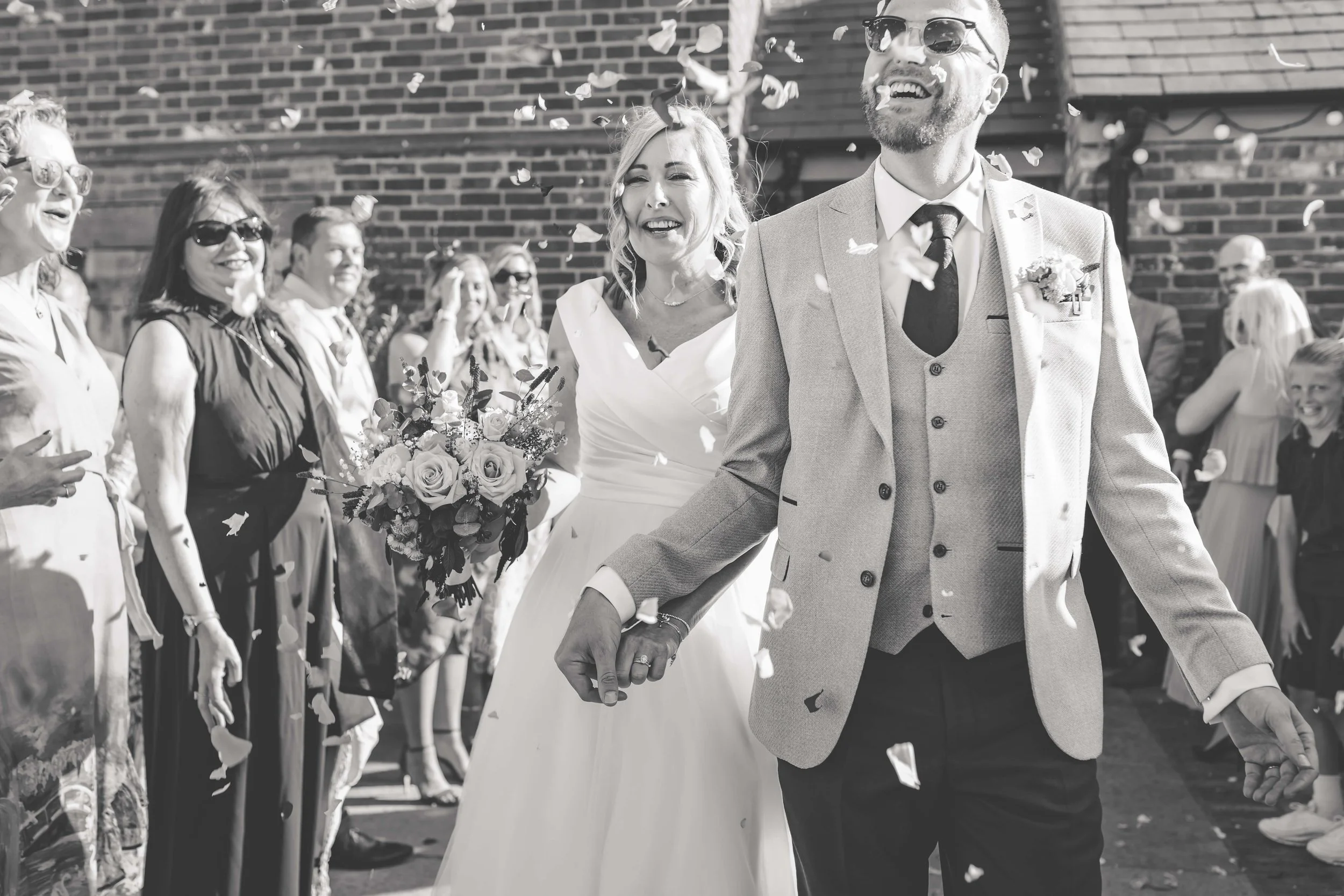 A black and white photo of a wedding moment with a smiling bride and groom holding hands, surrounded by guests throwing confetti, with a brick wall in the background.