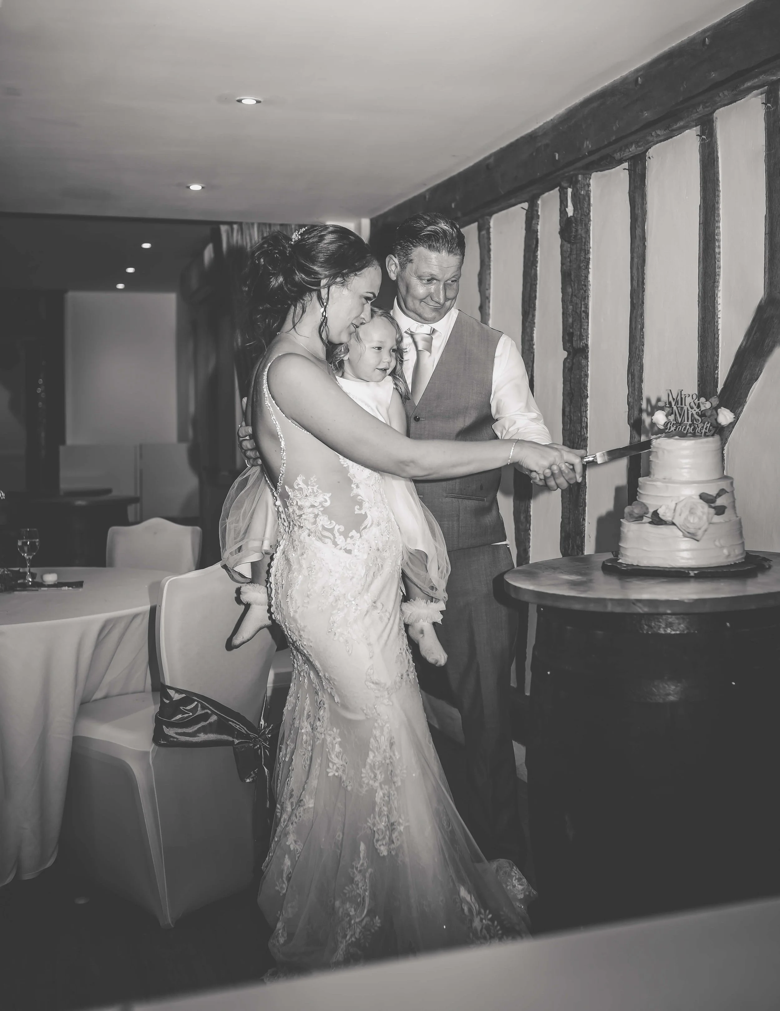 A black and white photograph of a wedding celebration where a bride and groom are cutting a wedding cake together. The bride holds a young girl, possibly their daughter, as they all smile around the cake.