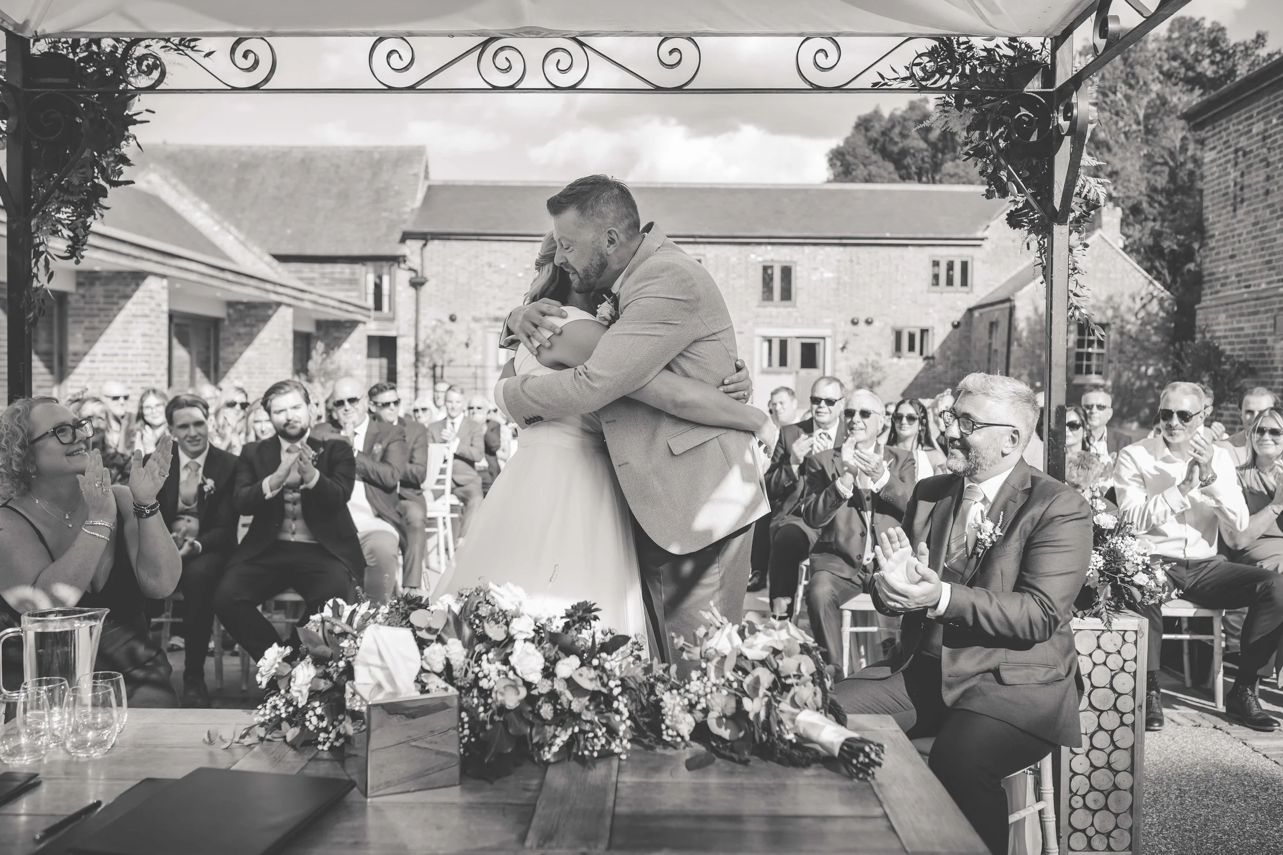 A wedding ceremony outdoors with a couple hugging at the altar, surrounded by seated guests clapping and smiling, under a decorative canopy, in a courtyard with brick buildings in the background.