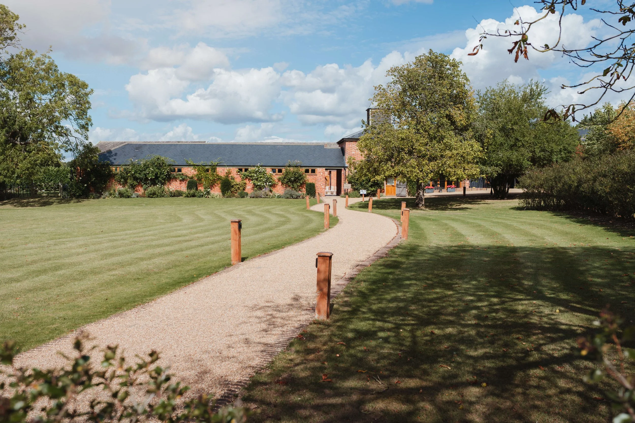 A gravel walking path curving through a well-maintained grassy park area with trees and bushes, leading to a brick building in the background during daytime with partly cloudy skies.