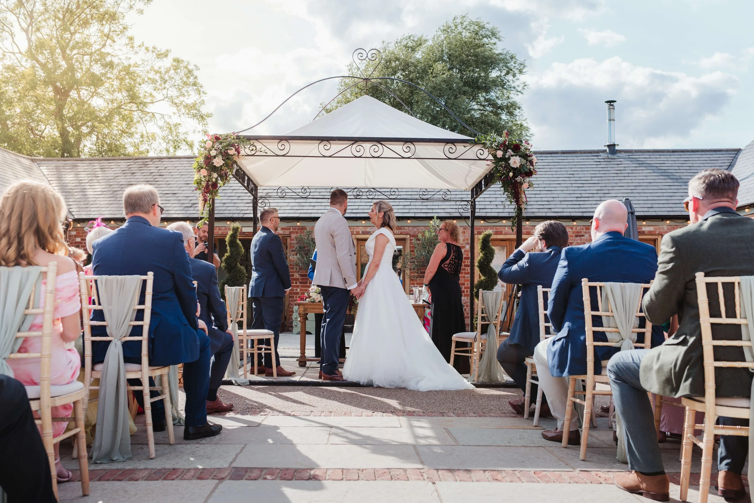 A wedding ceremony outdoors with the bride and groom holding hands at the altar, surrounded by seated guests, under a decorated canopy with flowers, on a sunny day.