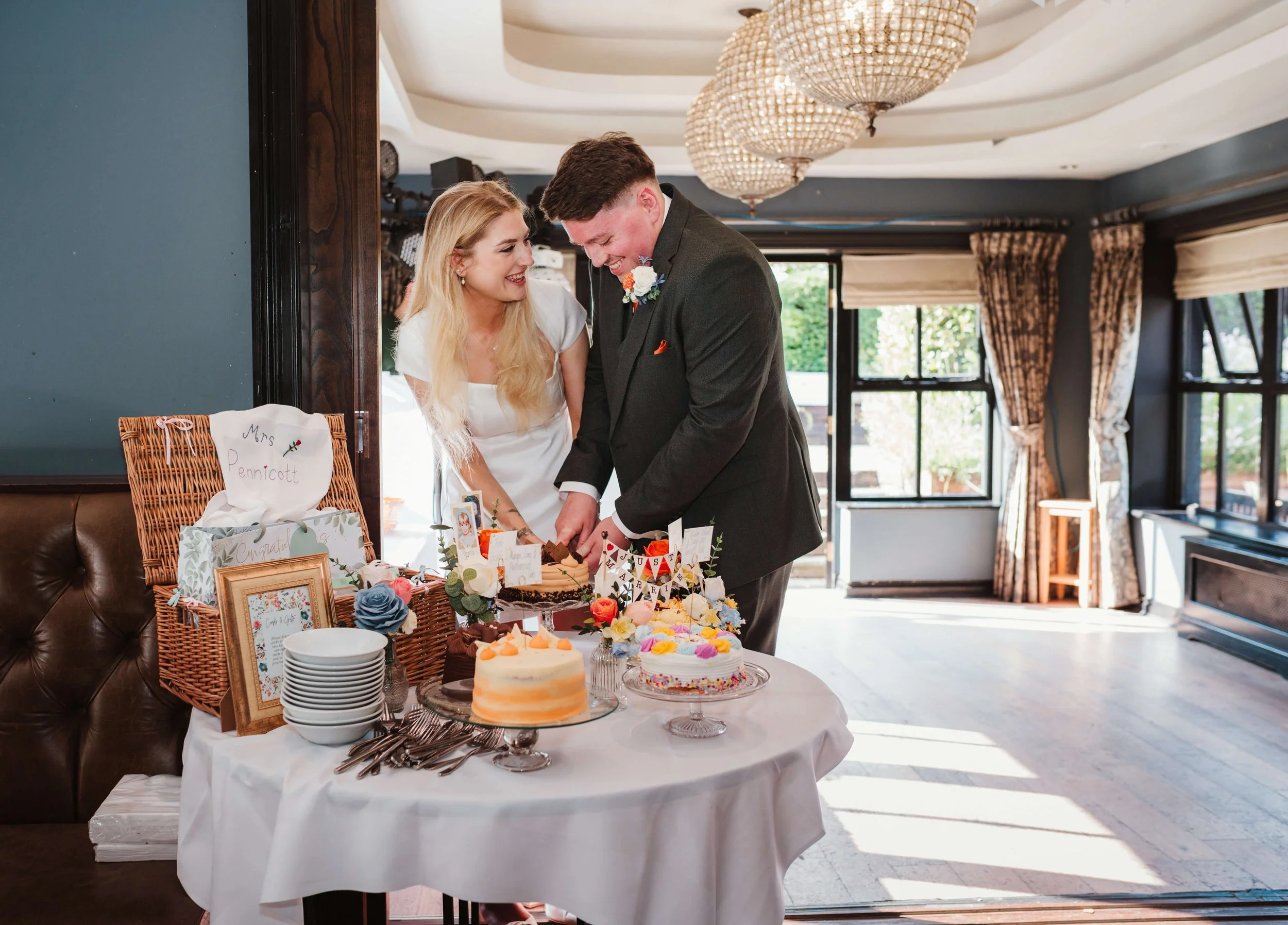 A bride and groom in wedding attire cutting their wedding cake at a reception, standing behind a table with decorated cakes, flowers, and gifts in a well-lit room with large windows and curtains.