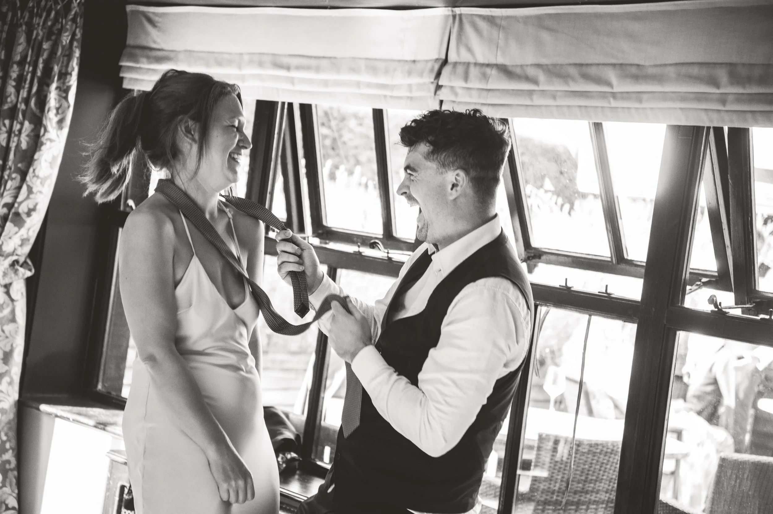 A man and woman laughing and smiling inside a vehicle, with the man holding a tie around the woman's neck.