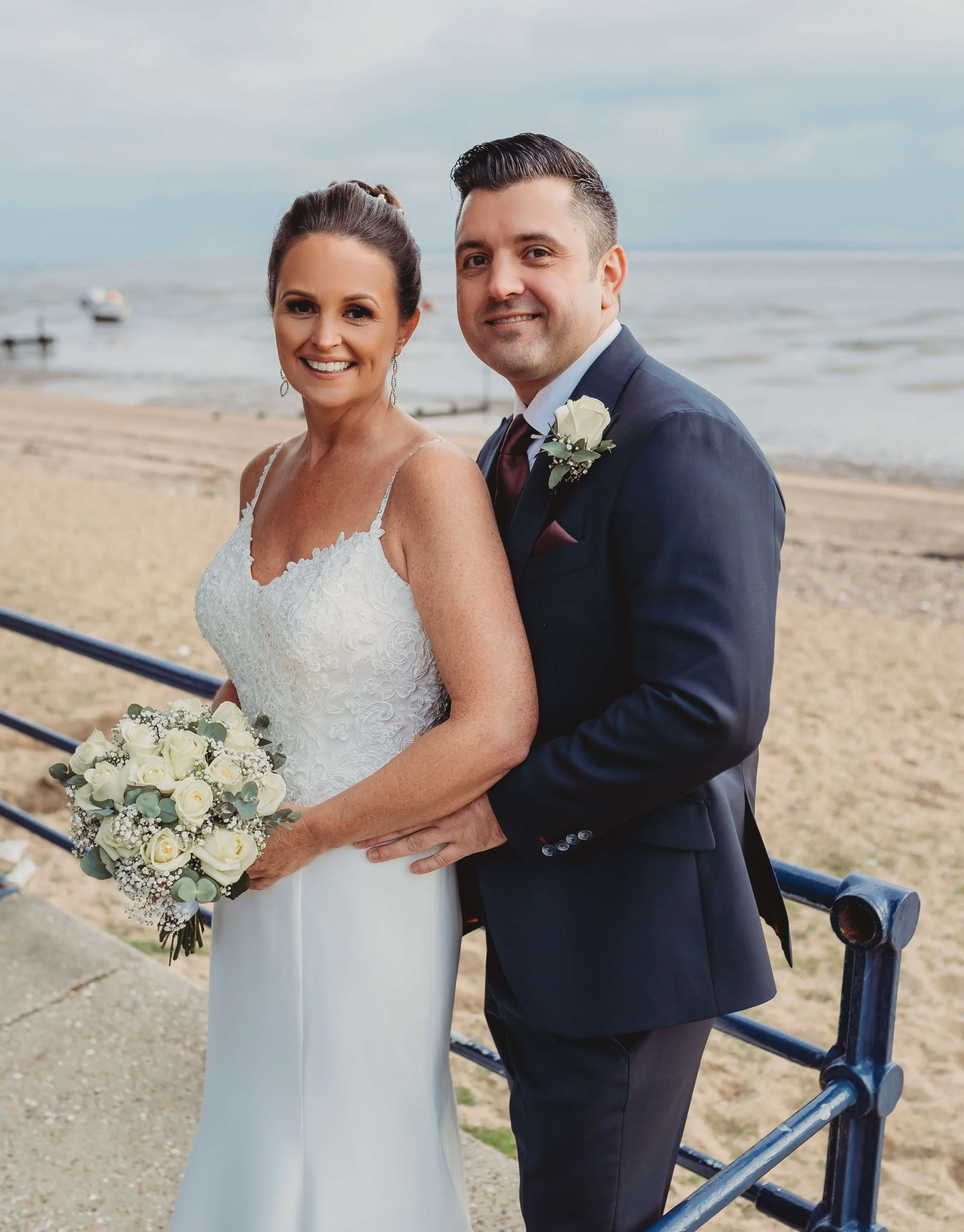 A newlywed couple in wedding attire posing on a beach front with ocean and boats in the background, smiling at the camera.