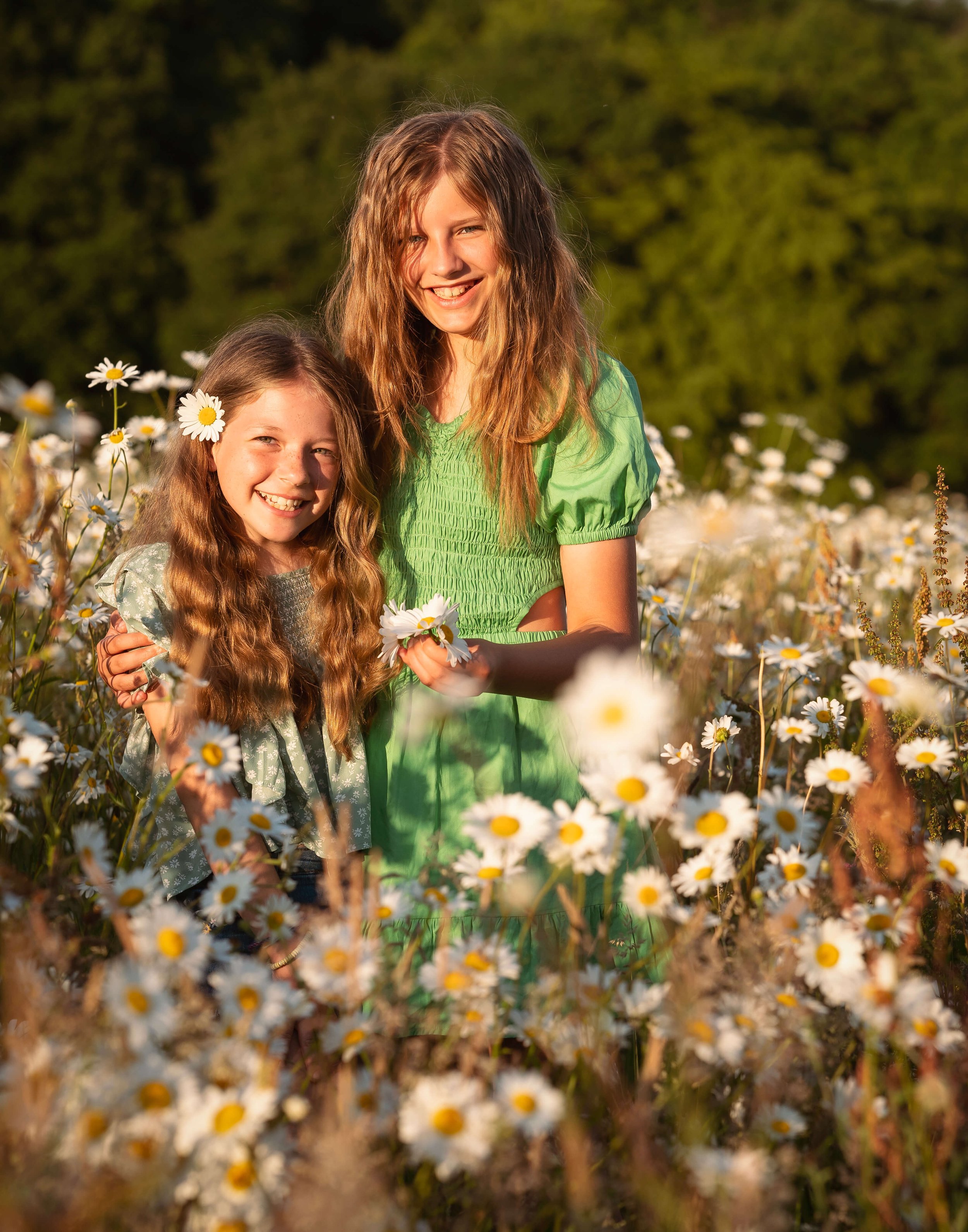Two girls with long, wavy hair smiling and standing in a field of daisies in the late afternoon sunlight.