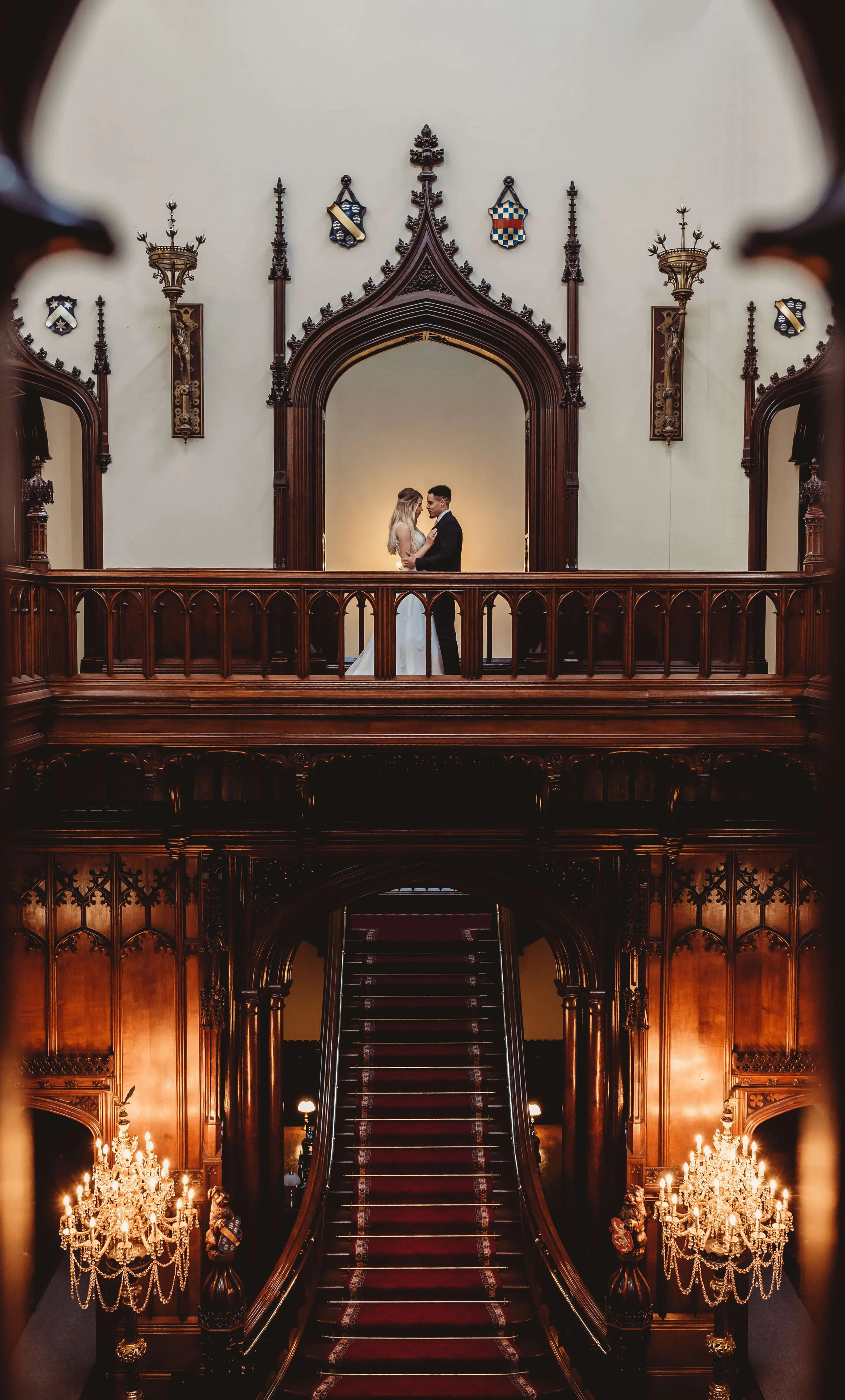 A bride and groom standing close together on an ornate wooden balcony inside a grand, historic building with intricate woodwork and chandeliers, captured through a framed opening.