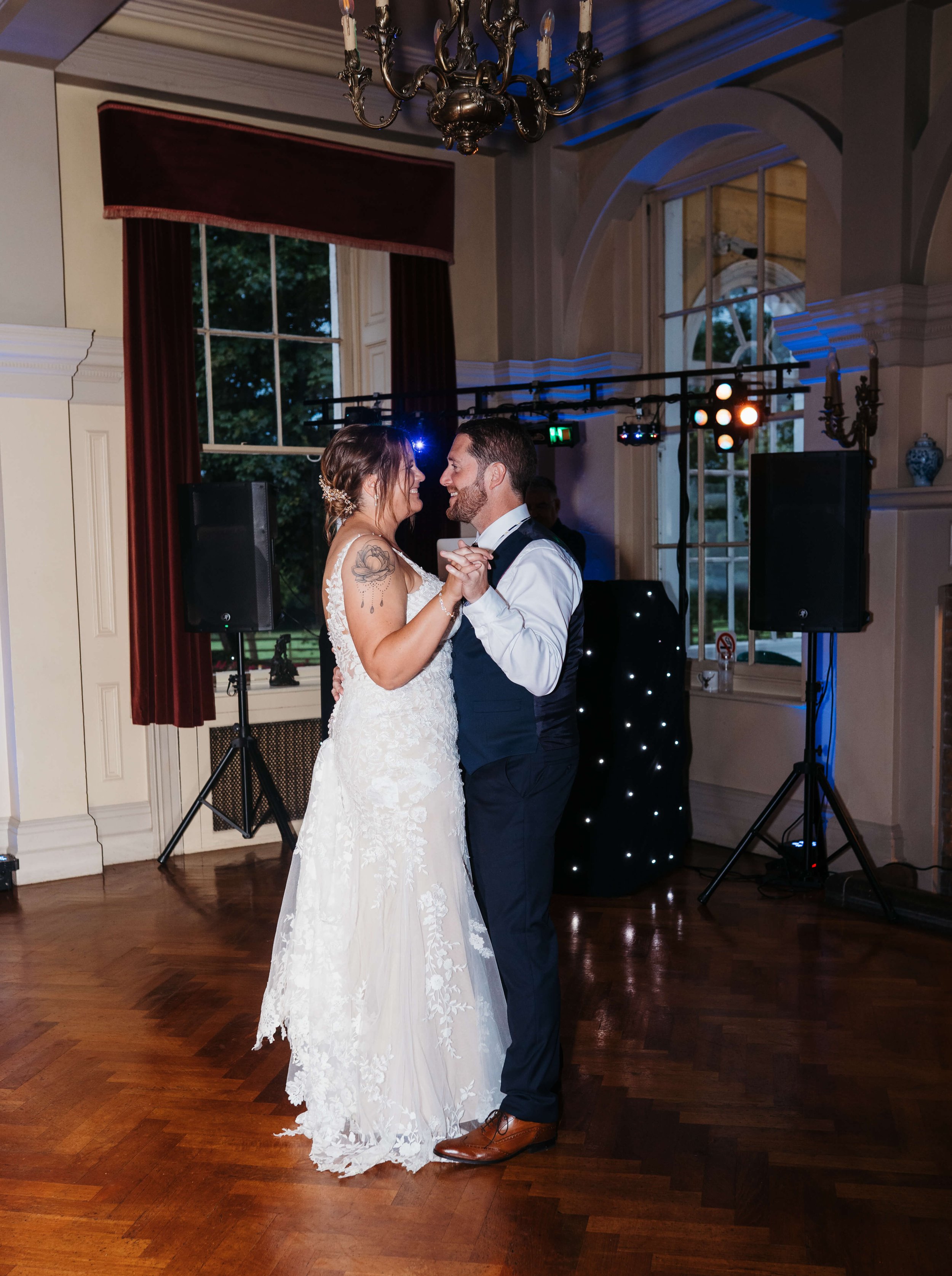 A bride and groom sharing their first dance at wedding reception in a decorated ballroom with large windows, red curtains, chandelier, and dance floor lighting.