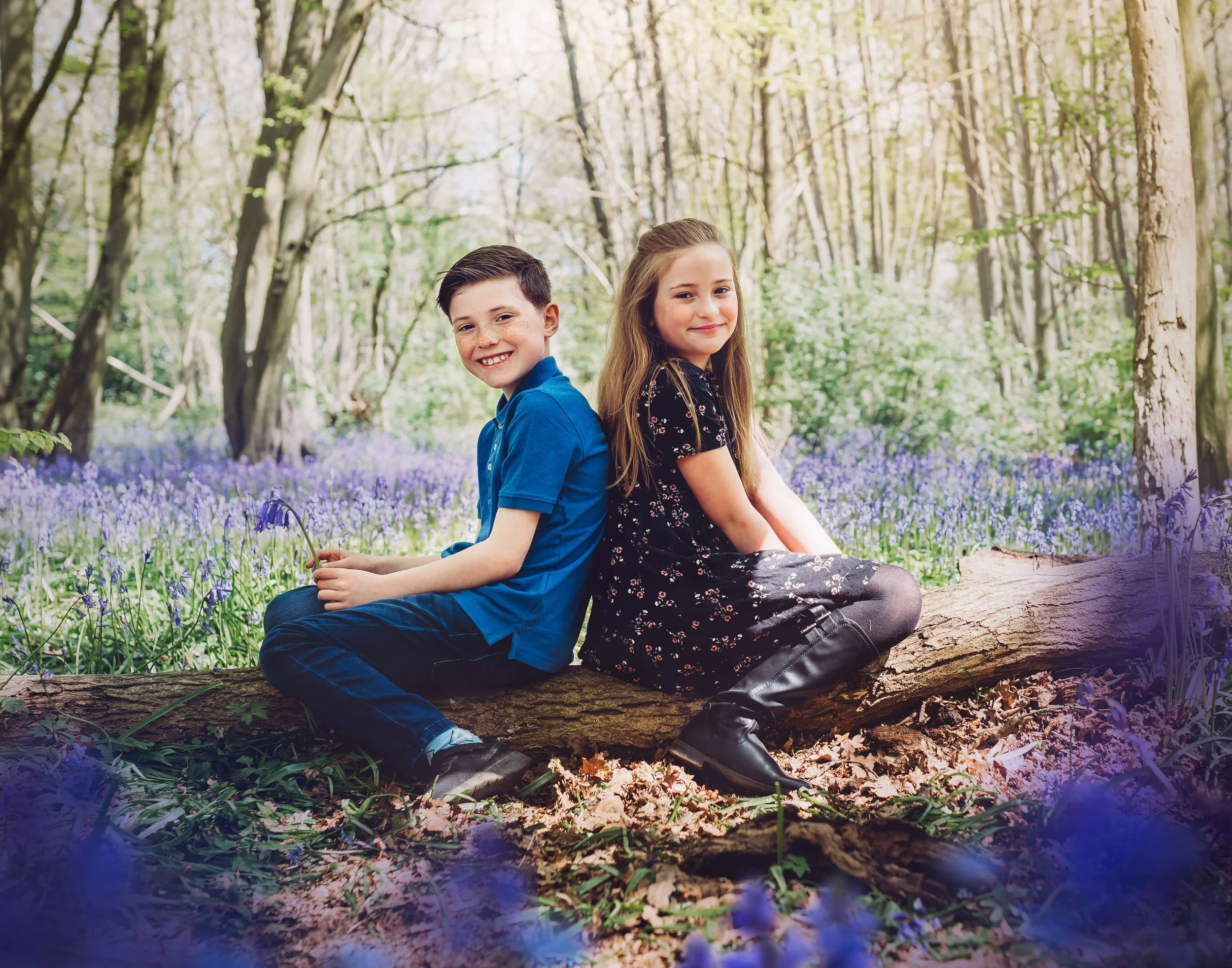 A boy and girl sitting back-to-back on a fallen log in a forest, surrounded by purple flowers and trees.