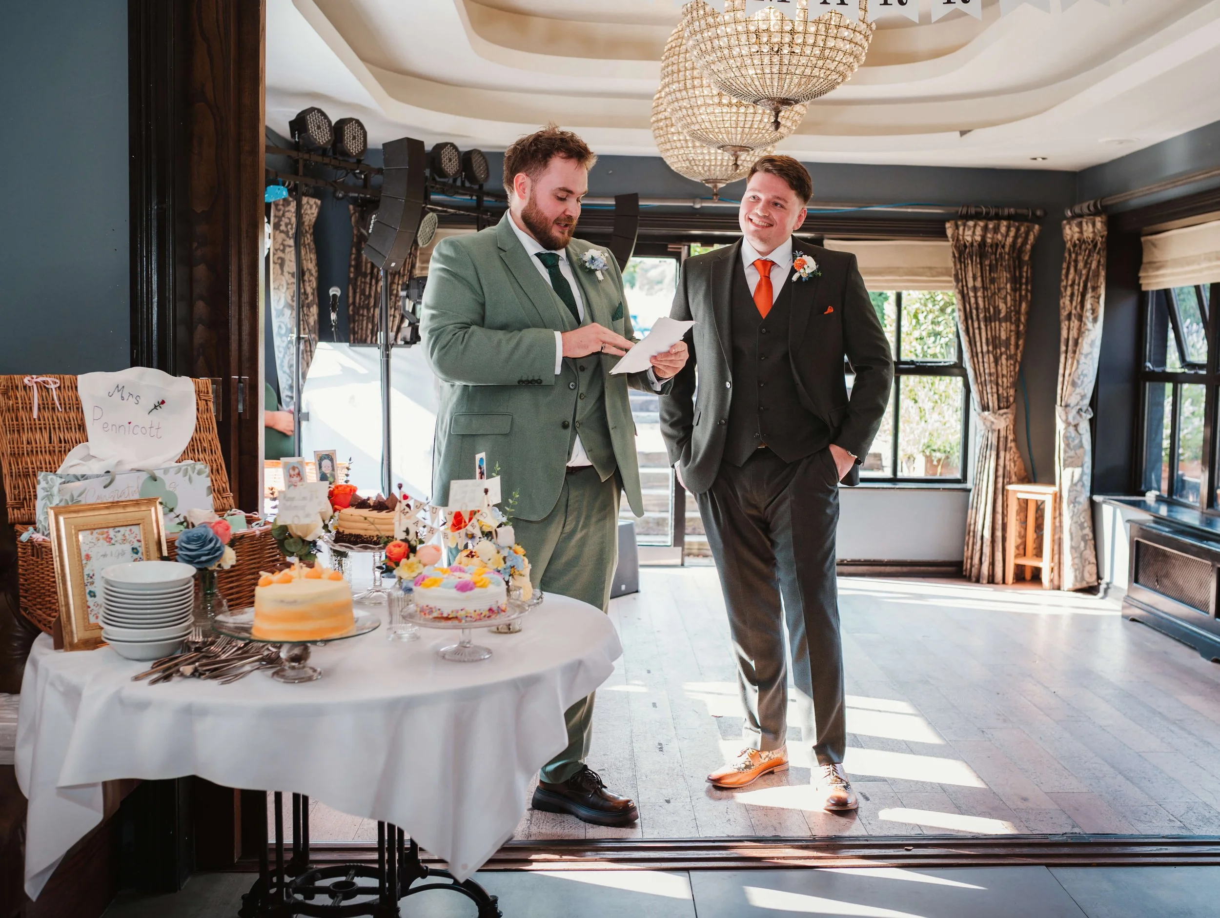 Two men in formal suits stand near a decorated table with cakes and small framed photos at an indoor wedding reception, smiling and engaging in conversation.