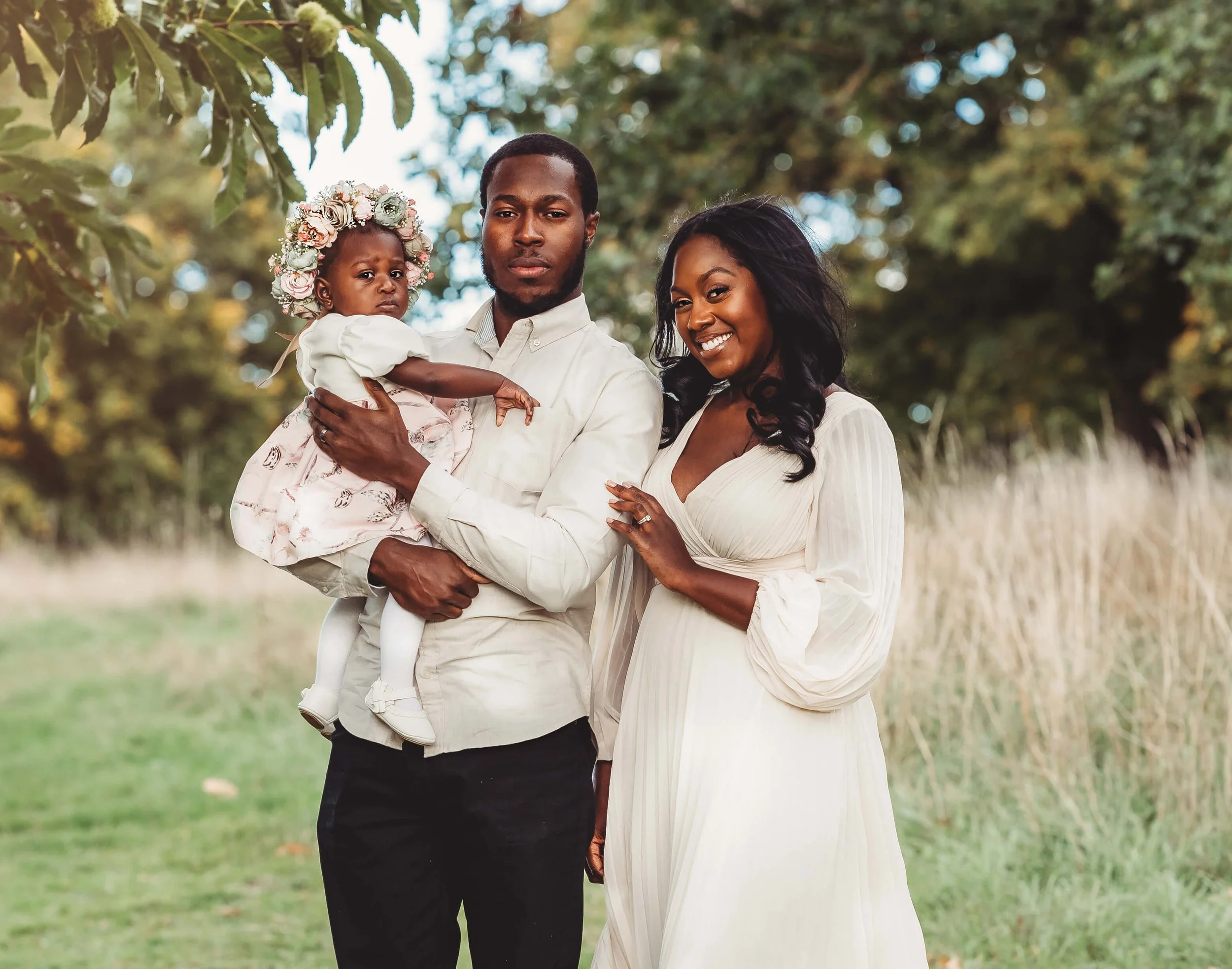 A Black family of three outdoors, with a man holding a young girl wearing a floral crown, and a woman in a white dress standing beside them, all smiling.