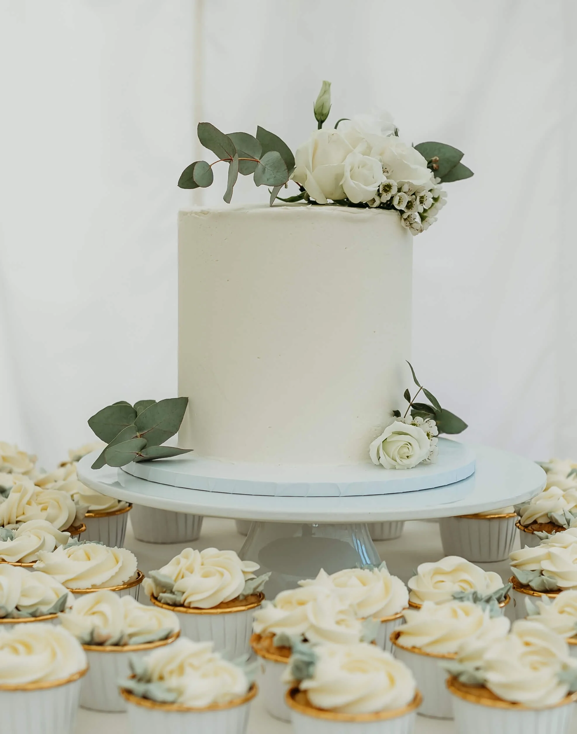 A white wedding cake decorated with white flowers and eucalyptus on a cake stand, surrounded by cupcakes with white frosting and eucalyptus decorations.