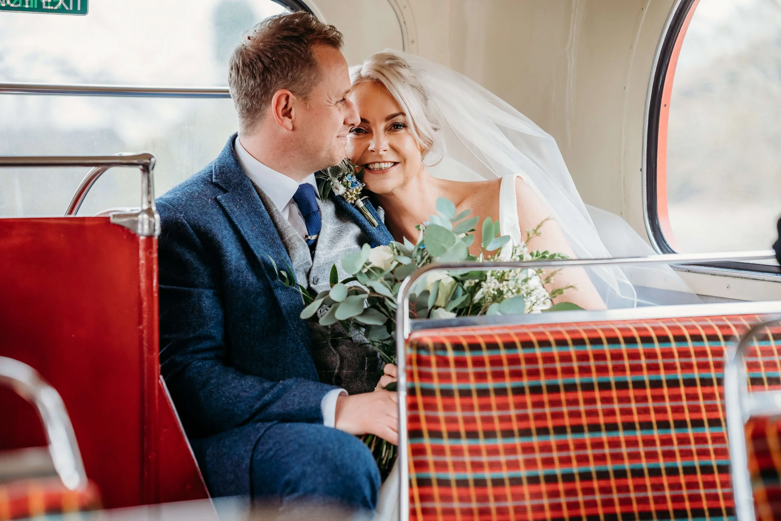 Bride and groom sitting on a bus, smiling and looking at each other, with the bride holding a bouquet of greenery and white flowers, and both dressed in wedding attire.