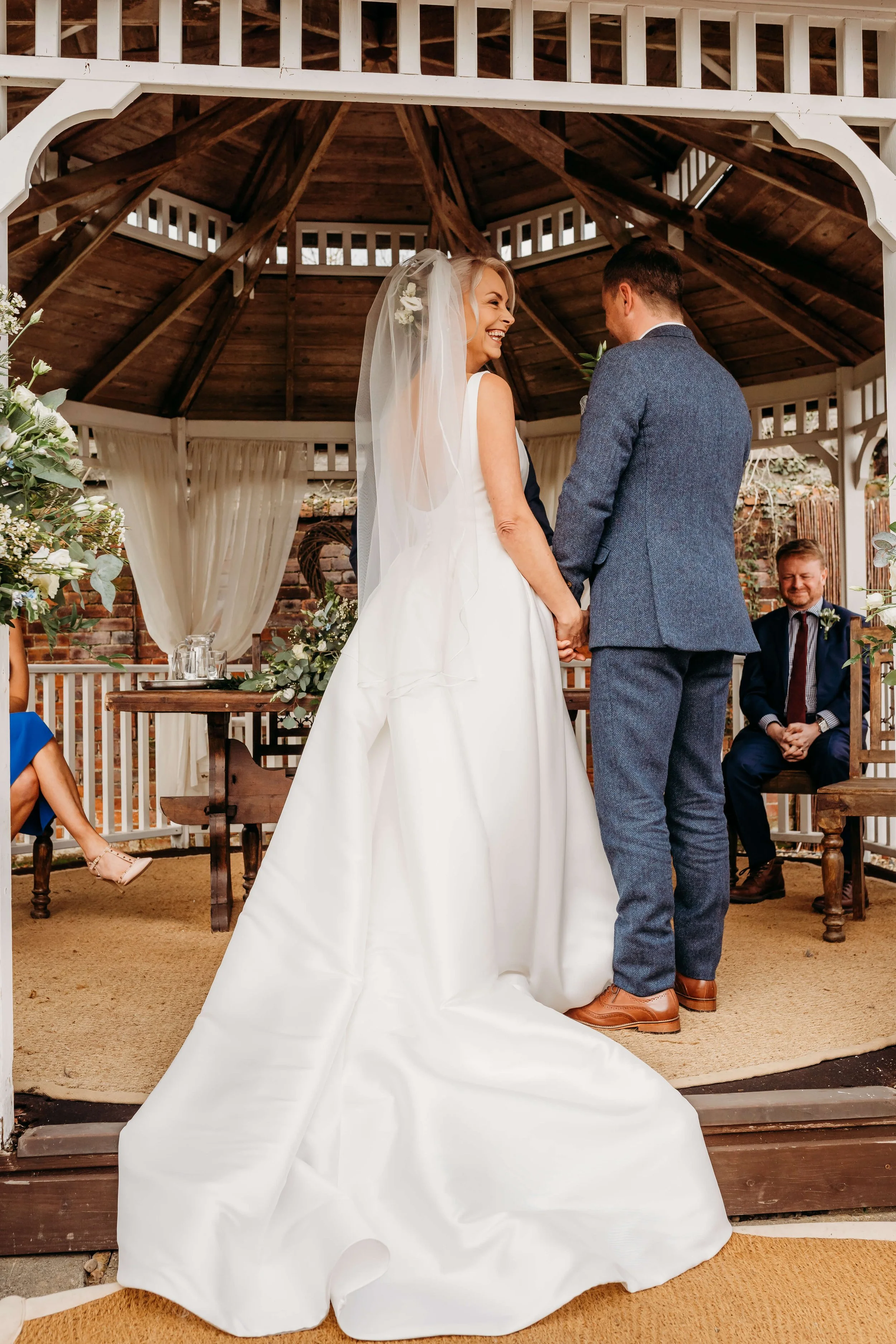 A bride and groom holding hands during their wedding ceremony inside a wooden gazebo, with a smiling man seated in the background.