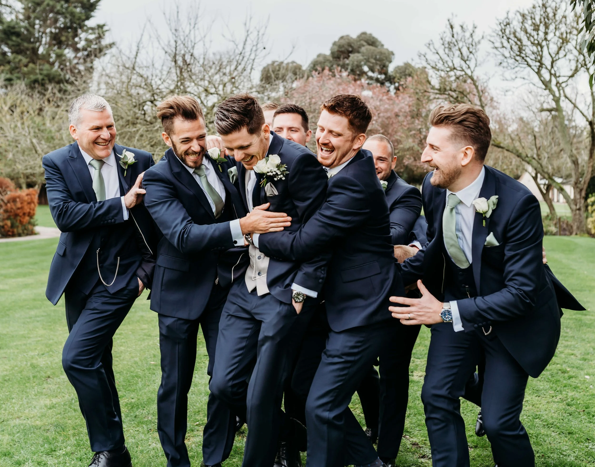 Group of men in suits laughing and playfully pushing each other in a park on a cloudy day.