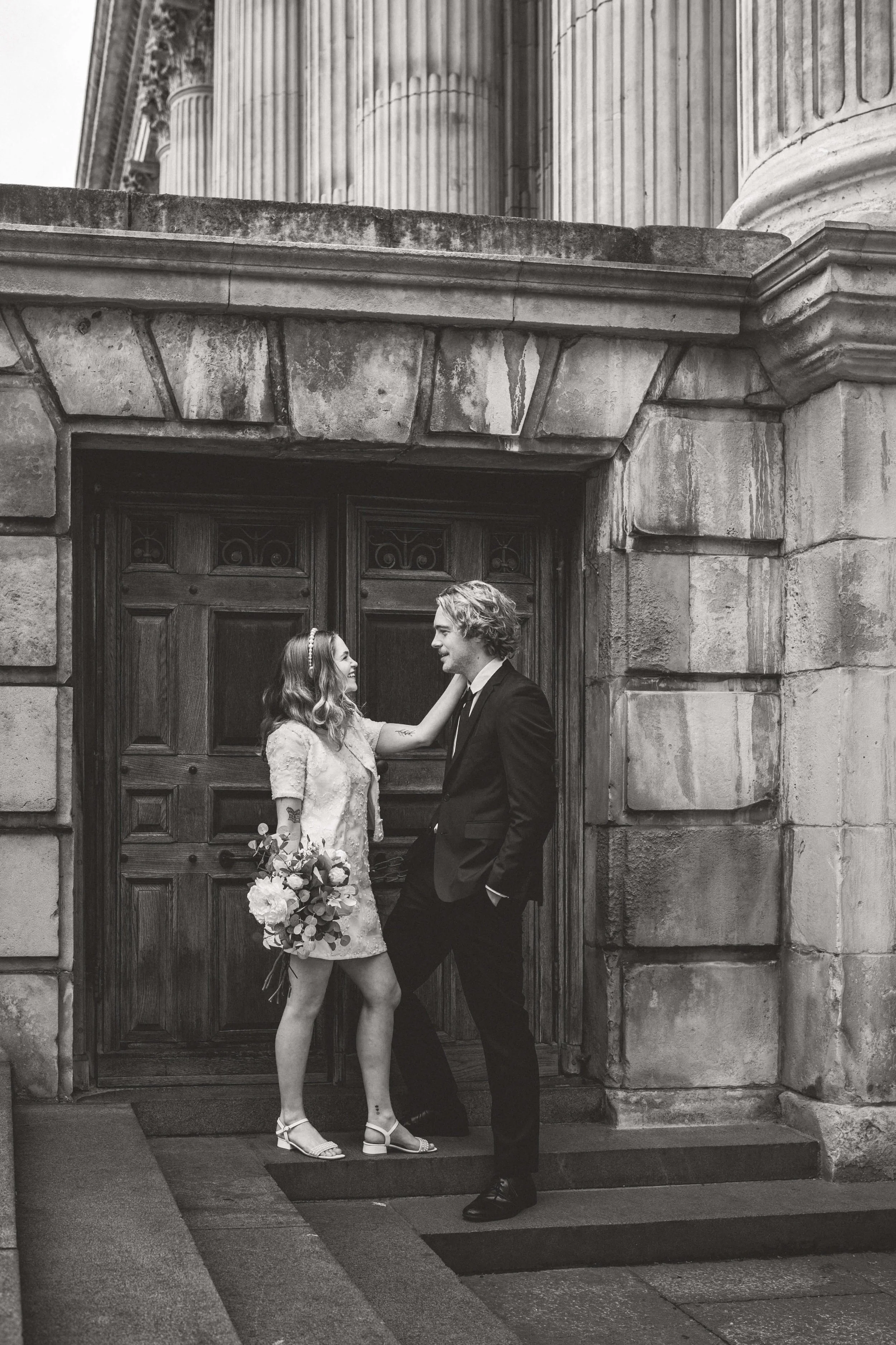 A black-and-white photo of a couple outside a historic building, with the woman wearing a dress and holding a bouquet, and the man in a suit, engaging in a romantic moment.
