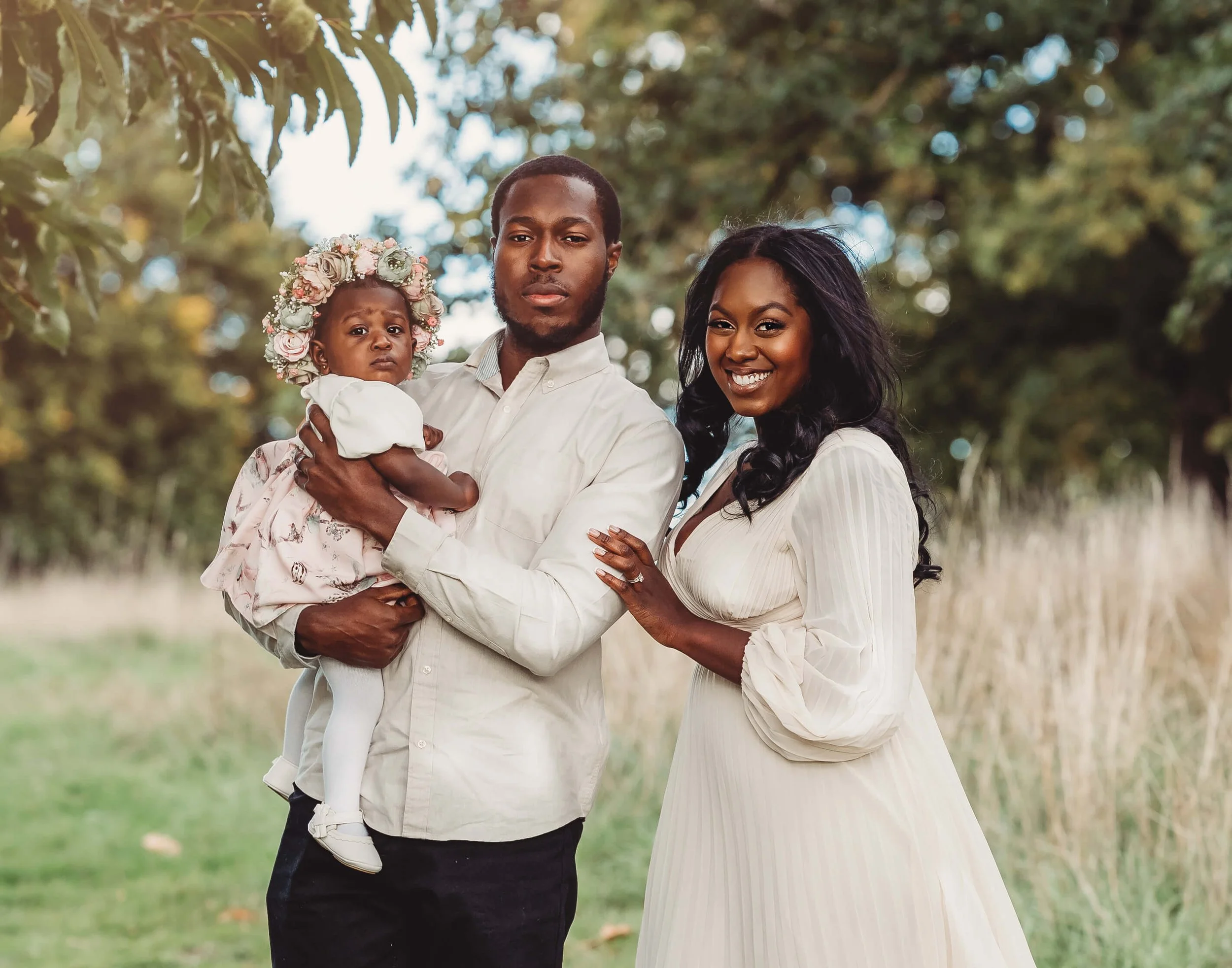 A happy family of three standing outdoors in a park during the daytime. The father, holding a young girl wearing a floral crown, is standing next to the smiling mother. The scene is surrounded by trees and nature.